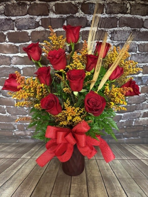 Red roses arranged in a tall vase with yellow filler and a red bow.