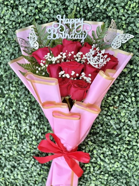 Pink Congratulations Flat Bouquet of Roses - A tight cluster of vivid red roses wrapped in soft pink paper with gold edging creates an instantly celebratory, romantic statement - perfect as a graduation bouquet or a birthday roses gift. The gold "Congrats Grad" and other celebratory toppers plus decorative butterfly picks make the arrangement read unmistakably milestone-ready, so the person you're celebrating will feel seen and special the moment they open it.  The bouquet pairs richly colored red roses with small white filler blooms and fern-like greenery for contrast and texture. Wrapped in pink paper, finished with a bright red ribbon and gold accents, it has a polished, gift-like presentation that reads both youthful and elegant. Sized as a hand-tied flat bouquet, it sits comfortably in the hand and is easy to carry to ceremonies or surprise visits.  Choose this arrangement for graduations, birthdays, anniversaries, or any moment you want to mark with warmth and flair - the toppers make the occasion clear while the classic roses convey affection. For best enjoyment, keep the stems in fresh water, trim them slightly before placing in a vase, and display out of direct sunlight to extend vase life.