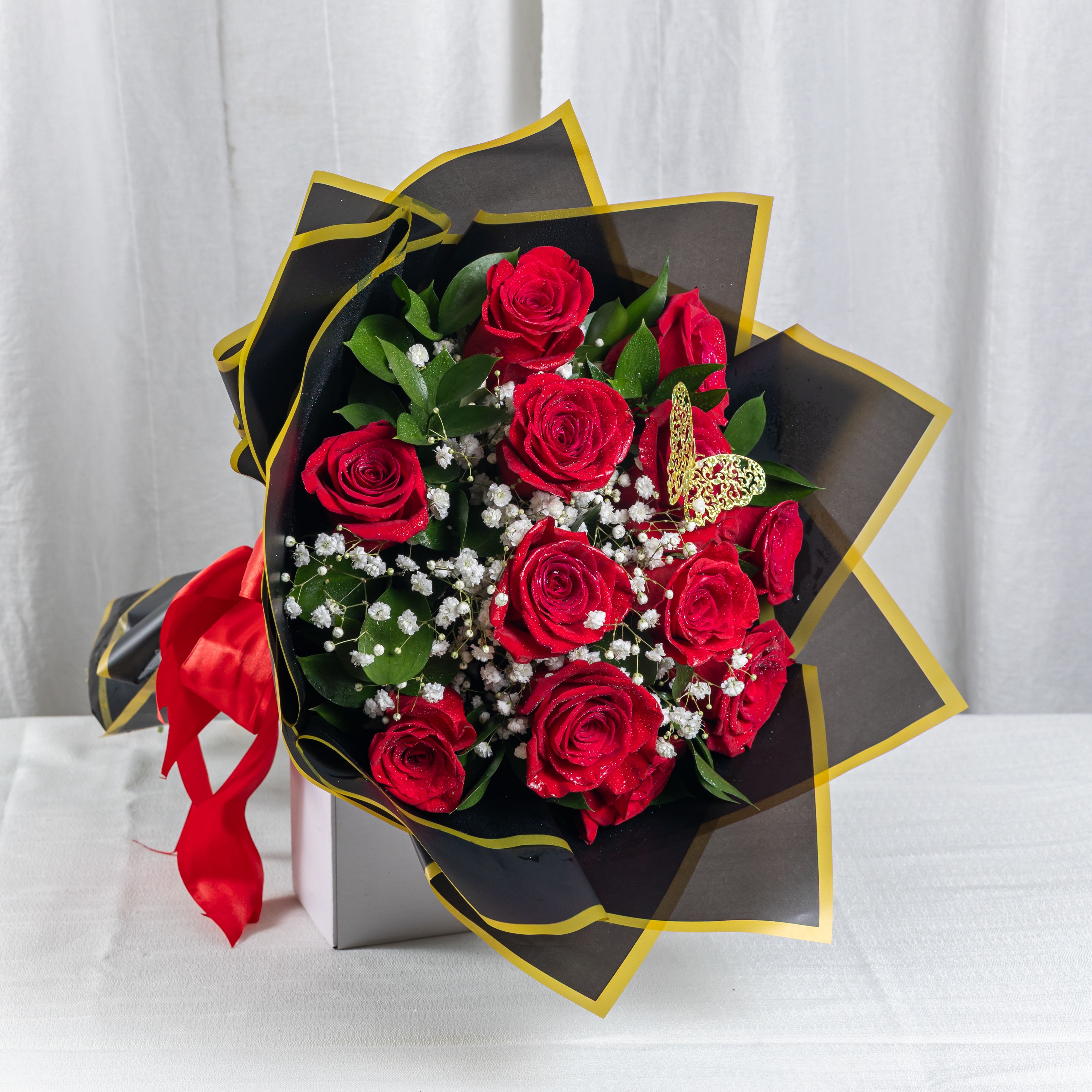 Velvet Devotion (12 Roses) - A stunning bouquet of 12 red roses wrapped in black and gold-trimmed paper, accented with baby’s breath and lush greenery. Nestled in a silver box and tied with a bold red satin ribbon, this arrangement speaks of passion, elegance, and timeless romance.