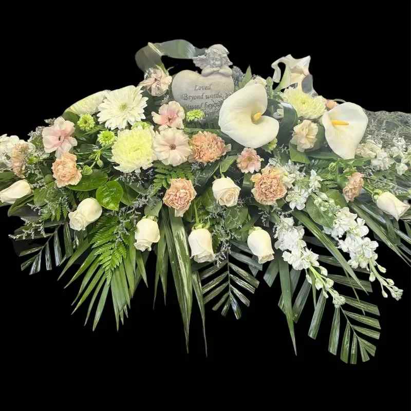 Angelica - A serene sympathy spray crafted to comfort and console, this flower arrangement is ideal for funeral or memorial services. The large, low design centers on elegant white calla lilies and soft blush roses to convey reverence, while the mix of carnations, daisy-like chrysanthemums and clustered filler flowers adds gentle texture-perfect for honoring the person you're thinking of.  Layered over broad palm fronds and mixed greenery, the white, cream and pink palette reads calm and formal without feeling cold. A memorial heart plaque and a small angel figurine sit near the center to reinforce the remembrance message and provide a keepsake focal point. The low, horizontal silhouette makes it suitable for display on a casket or memorial table and ensures the flowers read beautifully from all sides.  Choose this arrangement when you want the recipient to feel remembered and comforted during a difficult time; it sends a message of sympathy and gentle support. Standard care for sympathy sprays is minimal once placed at the service-ask about delivery timing to ensure the arrangement arrives fresh for the ceremony.
