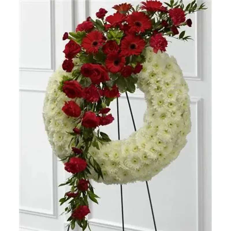 FTD® Graceful Tribute Wreath™ - Red roses, carnations and gerbera daisies are accented with burgundy mini carnations and a variety of lush greens to create a sweeping arrangement that glides across the face of a snow white wreath consisting of chrysanthemums making this a remembrance for one who has touched your life like none other. Displayed on a wire easel. 