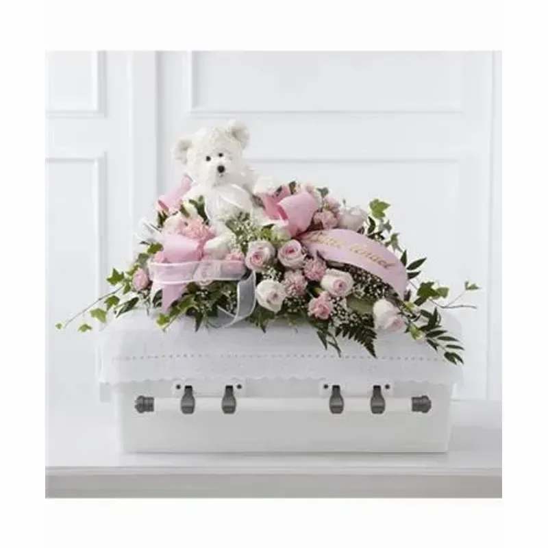 FTD® Touch of Sympathy Casket Spray™ - A tender spray of soft pink and white roses, carnations, daisies and airy filler designed as a sympathy casket spray for funeral and memorial services - the arrangement lays low and broad across a white casket to offer a gentle, comforting presence. The delicate palette and rounded blooms create a serene, consoling look that helps the person you're honoring feel remembered and cherished in a personal, heartfelt way.  This tribute includes clustered rose-like blooms, small carnations and daisies woven with baby's breath and trailing greenery for a layered, softened edge. Pale pink ribbons and a small white teddy bear nestled among the flowers add a sweet, affectionate detail, while the overall dimensions are intended to drape gracefully across the casket lid for a balanced, memorial presentation.  Appropriate for family tributes, services for a child or any loved one who appreciated soft, quiet florals, this spray communicates care and consolation without formality. Arrives ready to place on a white casket; stems are arranged for stability and easy placement - no extra assembly needed.