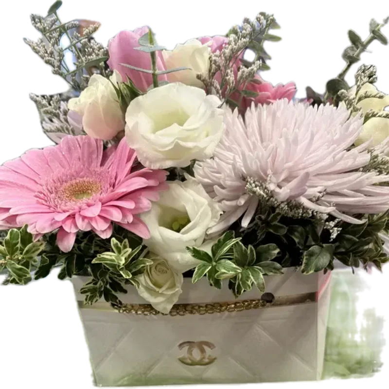 Luxury Designer Purse - A compact, gift-ready arrangement of soft pinks, creamy whites and pale lavender accents that reads like a luxury designer purse-perfect for an anniversary, birthday, or a "thinking of you" surprise. This flower arrangement for love and romance or a thoughtful thank-you uses a feminine palette and plush, rounded shapes to make the recipient feel cherished and seen; it also works beautifully as a get-well or just-because pick-me-up in spring and year-round.  Closely packed blooms include large pink gerbera daisies, pale chrysanthemum-style flowers, spray roses, lisianthus and tulip-like forms, with variegated foliage and airy filler adding texture around the edges. Everything is presented in a white decorative box trimmed in gold with a metal logo emblem and chain-style trim for a polished, high-end look. The compact size makes it ideal for bedside tables, desks, or a hallway console-an elegant, feminine gift that arrives ready to display.