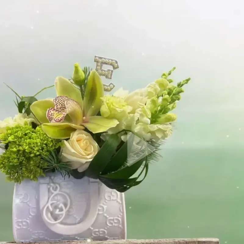 Gucci Inspired Floral - Lime-green orchids take center stage in this modern handbag-shaped floral arrangement, making a bold, fashion-forward statement perfect for a birthday, housewarming, or stylish thank-you gift. The fresh spring palette mixes vivid green blooms with soft white roses and snapdragons to create a lively contrast that reads contemporary and cheerful - the kind of bouquet that makes the person you're thinking of feel seen and celebrated.  Clustered hydrangea-like green blooms and upright white snapdragons add texture and height while pale-toned lilies, roses and hints of pink stock and other seasonal flowers soften the composition for a polished, approachable look. The arrangement arrives in a chic handbag container with a glittery letter pick tucked into the center for an extra touch of personality and presentation-ready flair.  Whether you're sending get-well wishes, a thoughtful thinking-of-you surprise, or congrats on a new home, this compact yet impactful design is easy to display and low-maintenance; swap the water every other day and trim stems as needed to keep blossoms fresh. It's an eye-catching gift that reads as both playful and refined - ideal for spring and wearable across the seasons.