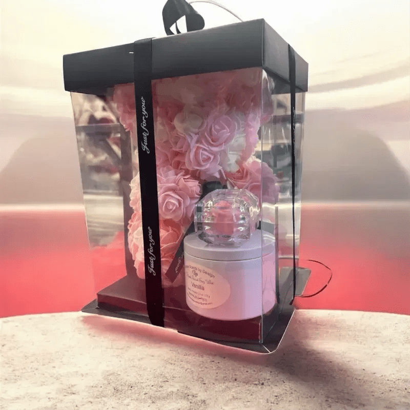 Diva Bear Pinky - A compact cluster of soft pink roses arranged inside a clear acrylic gift box creates a striking contrast with the black lid and ribbon-perfect as a romantic birthday bouquet, anniversary gift, or a thoughtful just-because surprise. Presented more like a curated gift set than a loose bouquet, the pink and white blooms read feminine and polished; the dark packaging gives the whole piece a modern, elegant edge that makes the recipient feel specially celebrated.  The arrangement includes a handful of pink roses (with white accents visible) seated neatly in the box, accompanied by a white candle jar and a small glass container at the base for an added touch of luxury and ritual. The clear acrylic box protects the blooms while letting their color shine, and the black ribbon finishes the look for easy gifting. Sized as a compact table-top arrangement, it arrives ready to display-no extra vase required-so the person you're thinking of can enjoy both the flowers and the little extras right away.