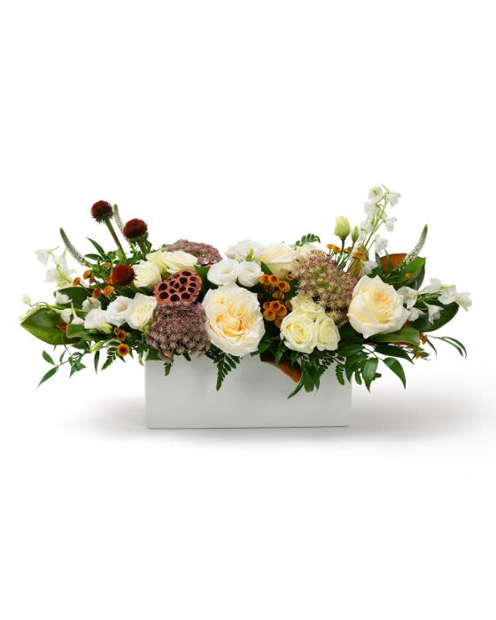 Low arrangement of cream roses with orange accents and seed pods in a white rectangular container
