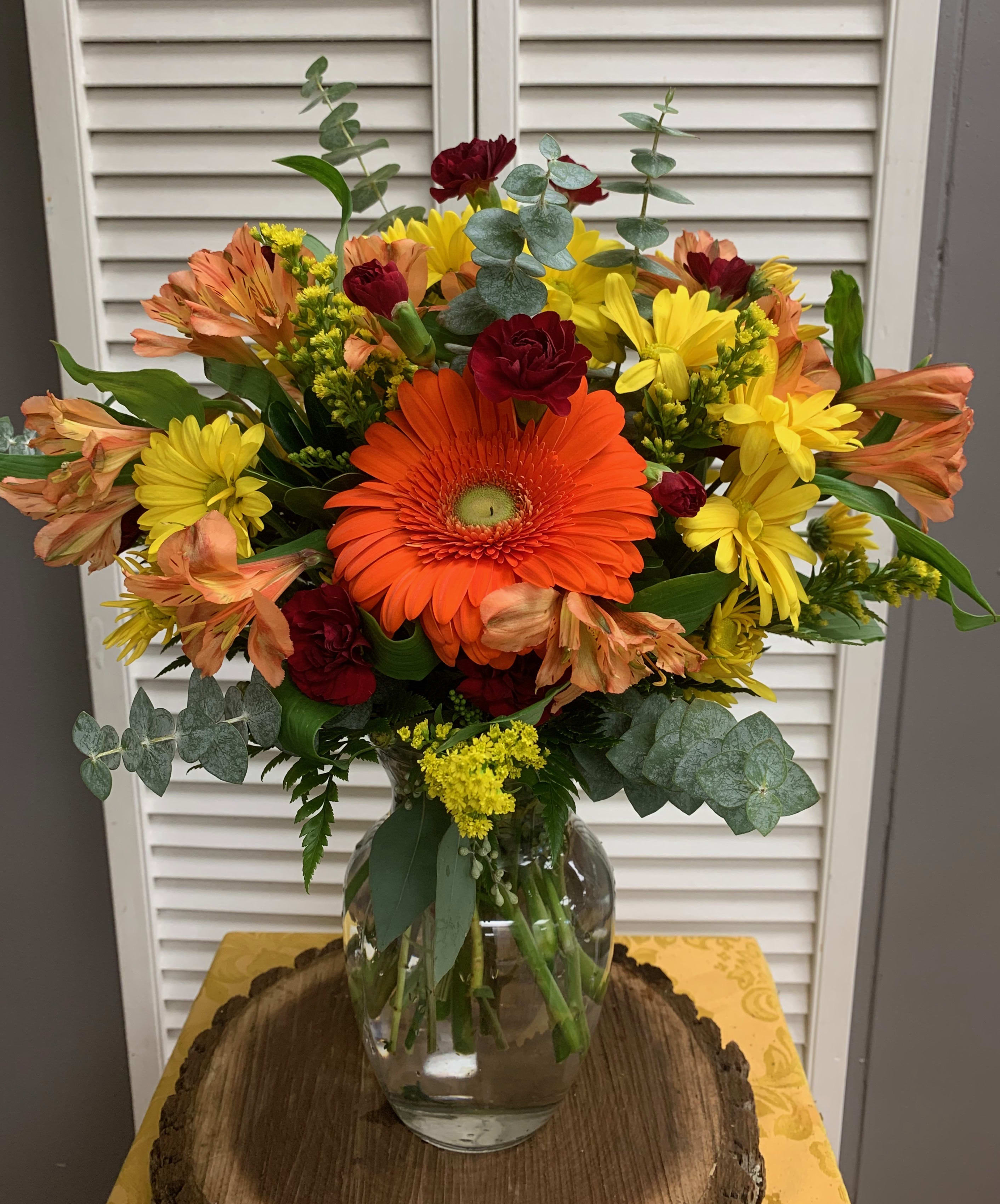 Mixed bouquet with orange gerbera daisies, yellow daisies, and red carnations in a glass vase