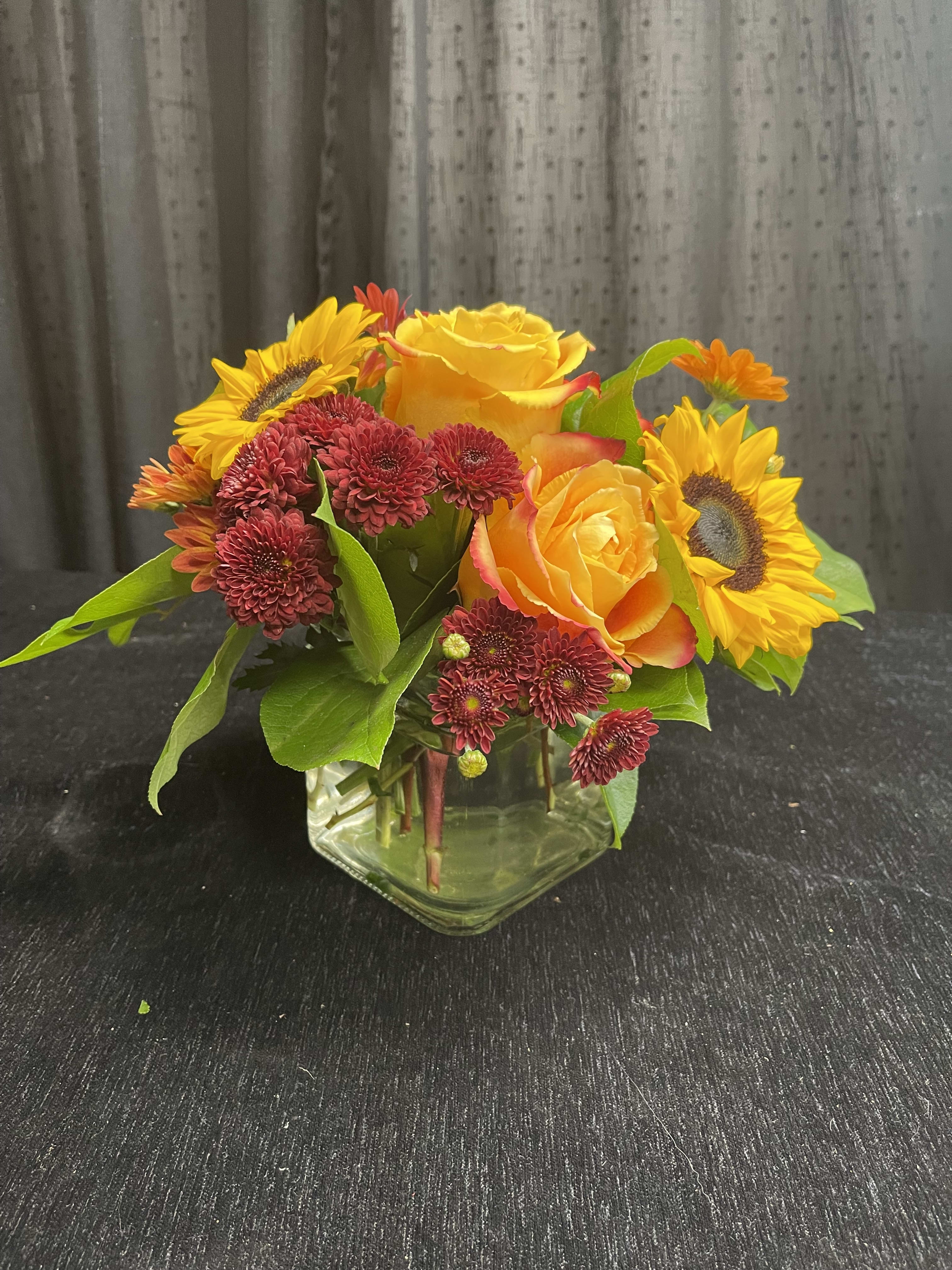 Sunflowers, yellow roses, and burgundy mums in a clear glass vase