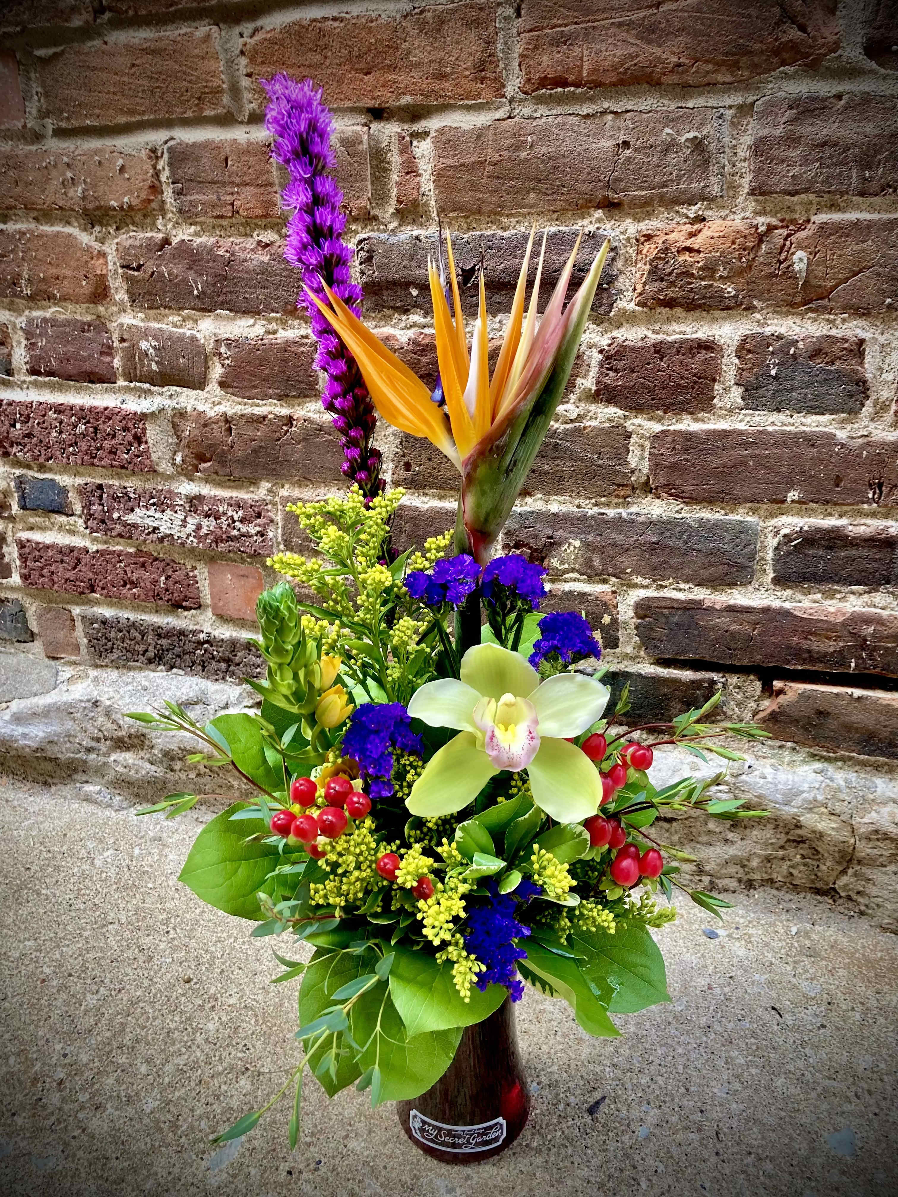 Sophistication  - A dramatic vertical silhouette anchored by a bird-of-paradise and pale green cymbidium orchid makes this tall flower arrangement an unforgettable Mother's Day gift that reads both exotic and elegant. The upright, sculptural line catches the eye while warm orange, yellow, magenta and purple blooms-set against glossy tropical foliage and long palm fronds-create a lush, layered presence that will make the person you're thinking of feel celebrated and noticed.  Clusters of vivid accent flowers and small red berry touches add lively contrast throughout the dense green base. Arranged in a decorative glass vase, the design balances one strong vertical bloom with a fuller lower cluster for stability and visual interest; the mix of orchids, tropical flowers and mixed greenery gives rich texture and long-lasting impact. Ideal for Mother's Day-this piece brings exotic color and a sophisticated flair to any room.