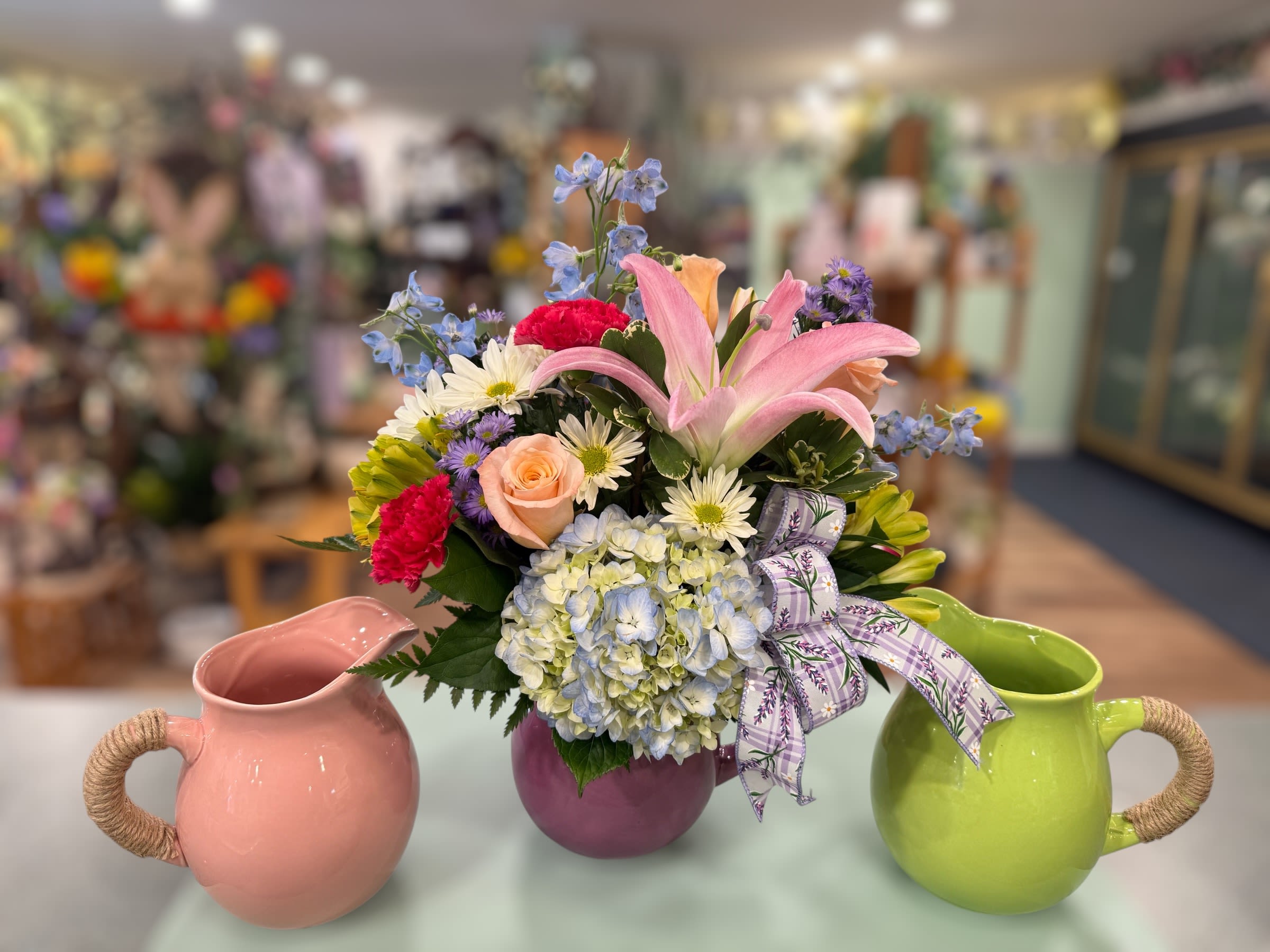 Garden Pitcher (GP) - Full of color and perfect for all occasions! Designed in a ceramic pitcher with a floral bow.  Pitcher color will vary between the three colors pictured (Purple, green or pink).  ***Flower color/type may vary depending on availability.