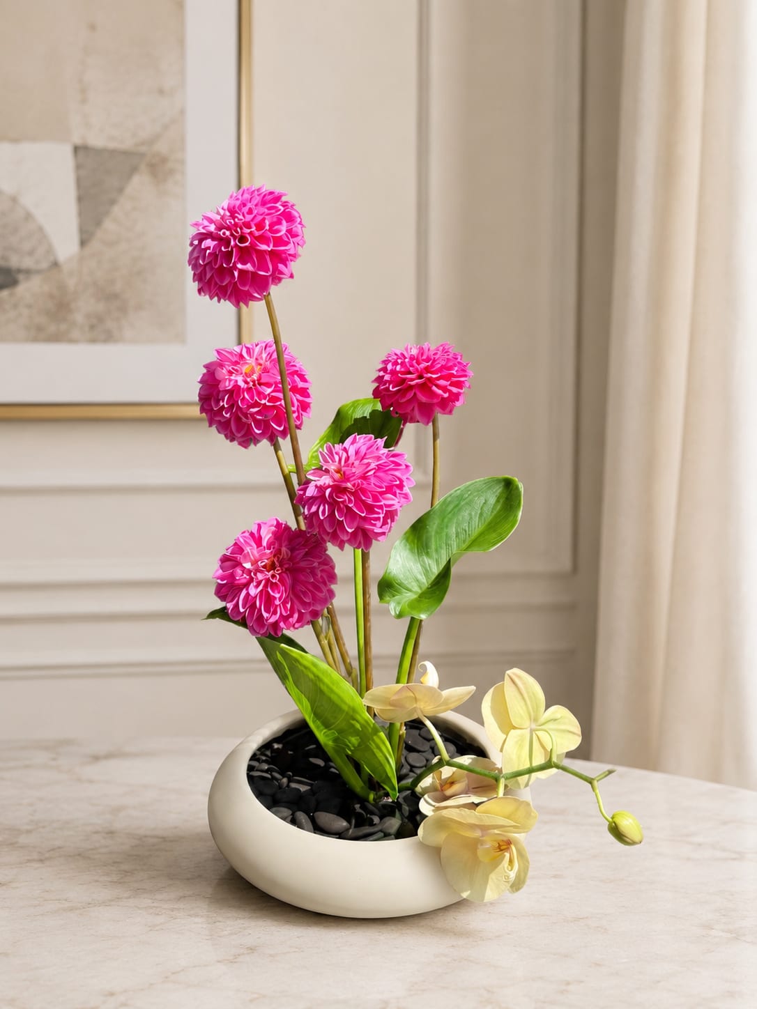 Dahlia Dreams - An elegant, modern, and vibrant floral arrangement designed in a white ceramic-style bowl with decorative black stones. The bright fuchsia dahlias bring bold color and energy, while the soft yellow orchids add a delicate and sophisticated touch. Tropical green leaves complete the design, creating a fresh, artistic piece perfect for any space.  Ideal for birthdays, thank-you gifts, special celebrations, or adding an elegant accent to a home or office.