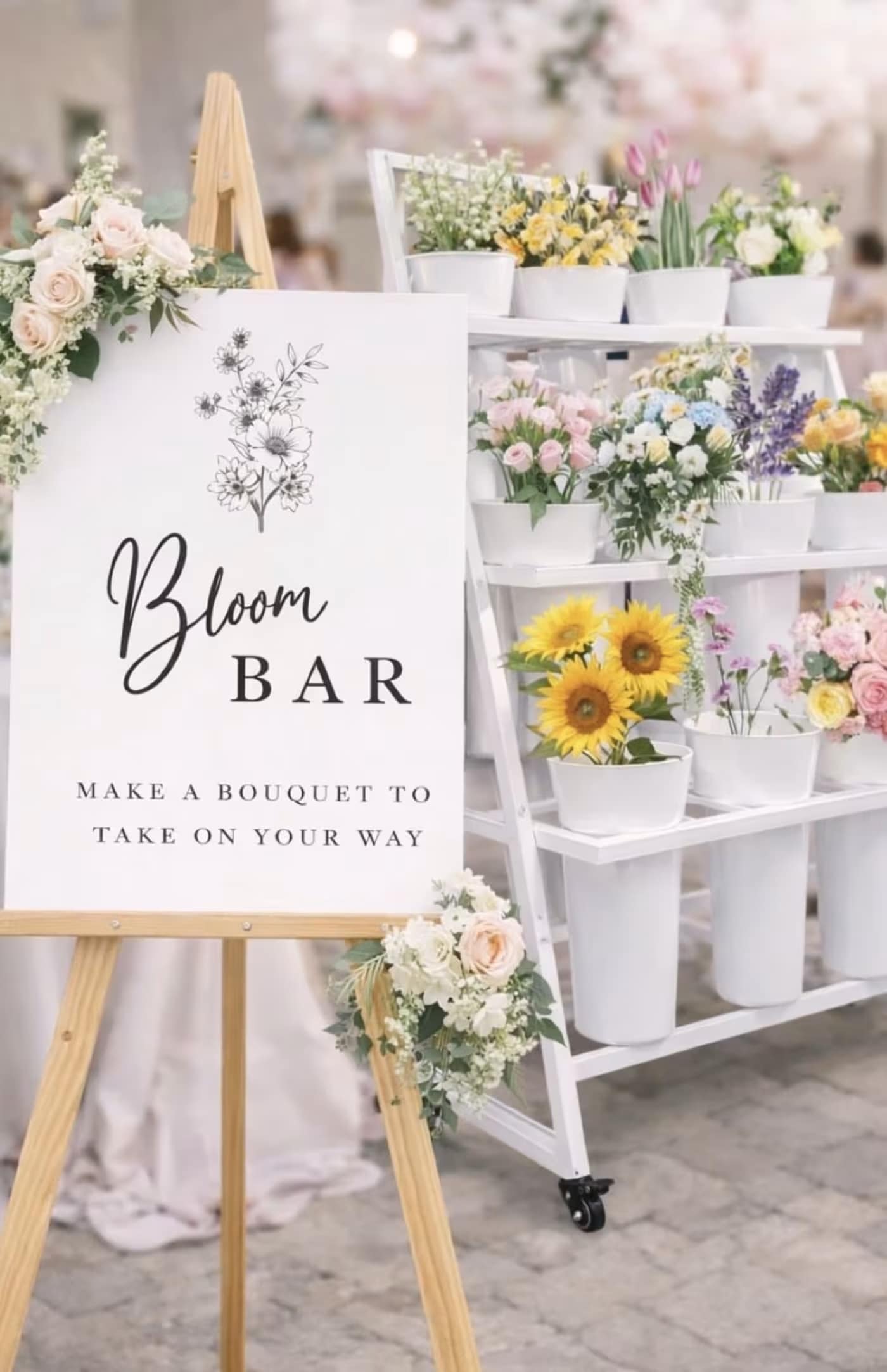 Bloom Bar by Town and Country Floral - Invite guests to create joyful, personal bouquets with a pastel, market-style bloom bar-perfect for weddings, bridal showers, baby showers, or any celebratory event. The setup centers on a large printed sign on a wooden easel and a clean white rolling cart stocked with white buckets, offering an airy, photo-ready station where the person you're celebrating can mix soft pink and white roses, pale tulips, purple accents, bright yellow gerbera daisies and sunflowers, and cheerful daisy stems.  Flowers arrive organized in separate buckets so guests can easily choose stems and assemble arrangements that feel handmade and thoughtful. The display is built around a white flower cart and matching buckets for a cohesive look; dimensions and exact stem counts are managed at booking to suit your guest count and event flow. This interactive centerpiece creates relaxed, memorable moments-book the bloom bar at least three days in advance to reserve the cart, easel, sign, and curated selection of blooms for your special day.