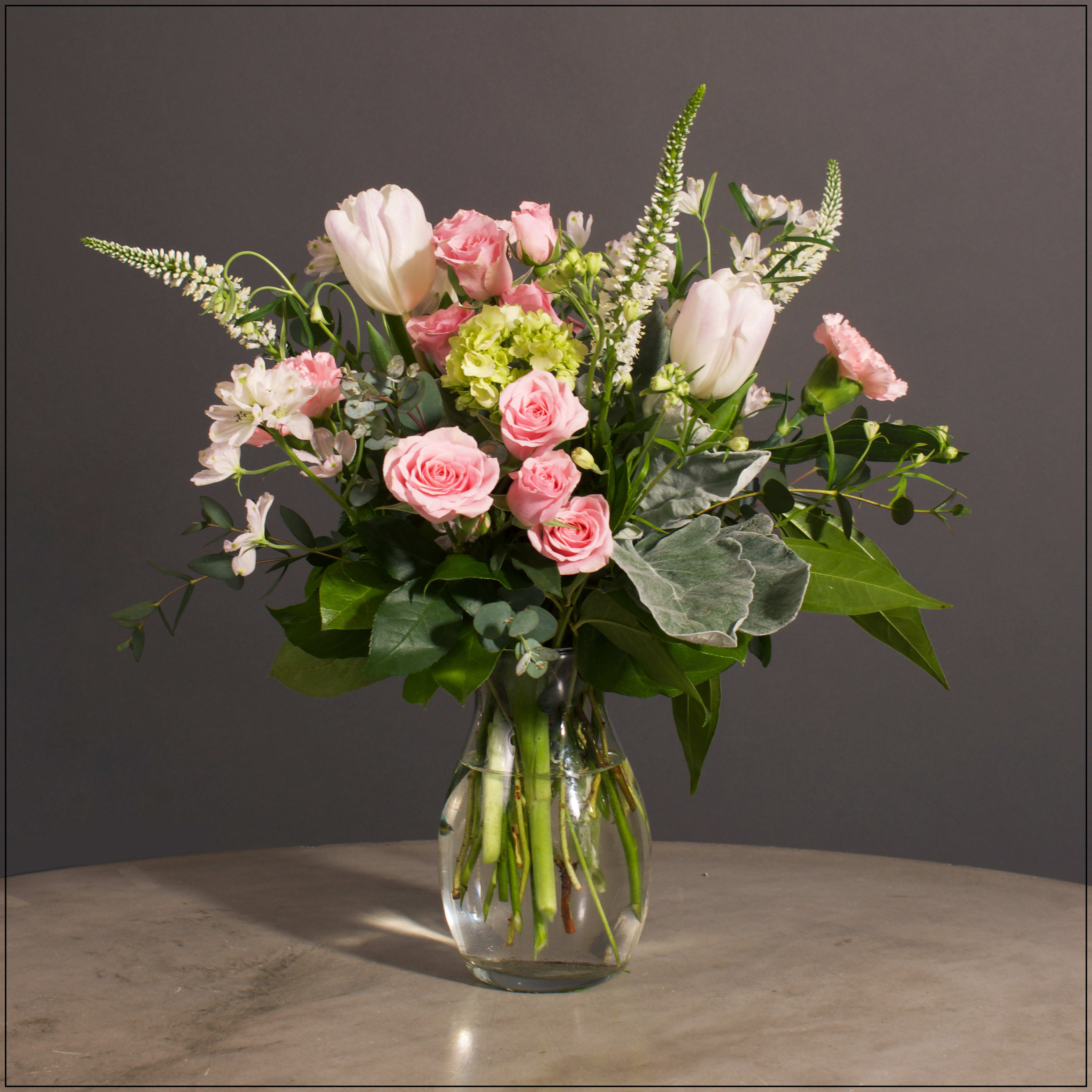 Lisbeth - Spray Roses, Carnations, Delphinium, Hydrangea and Veronica with Filler and Greens in a simple glass vase.  Approximately 17" H x 15" W