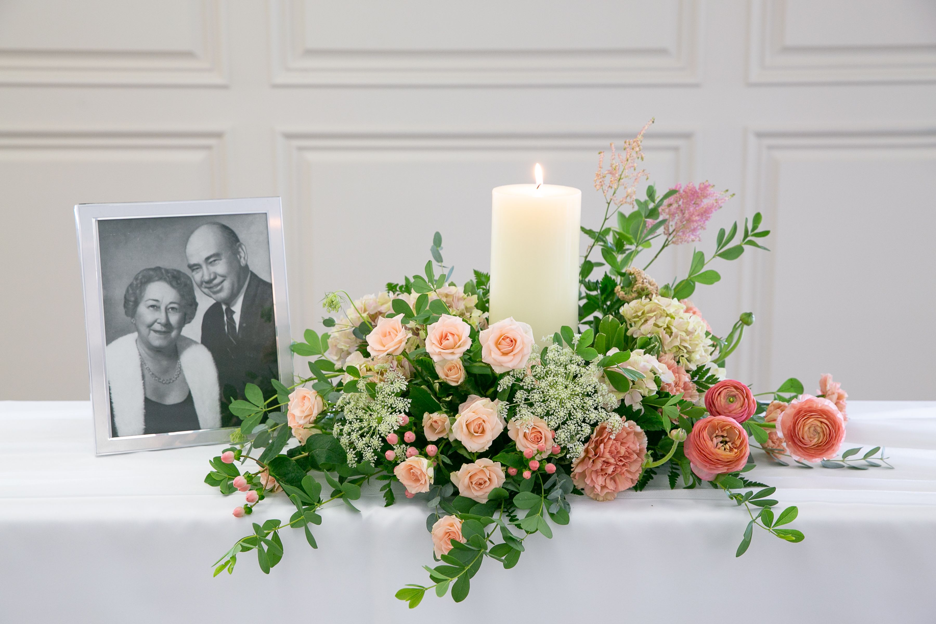 Forever Remembered™ Memorial Centerpiece - Create a meaningful tribute with our Forever Remembered™ Memorial Centerpiece, thoughtfully designed to honor a loved one's memory with grace and beauty.  A soft floral arrangement surrounds a premium pillar candle, creating a warm focal point for memorial services, celebration of life gatherings, or a remembrance table at home. Designed in delicate shades of blush, ivory, and soft pink with lush seasonal greenery, this centerpiece offers a peaceful expression of love and remembrance.  Frame and photograph are not included.
