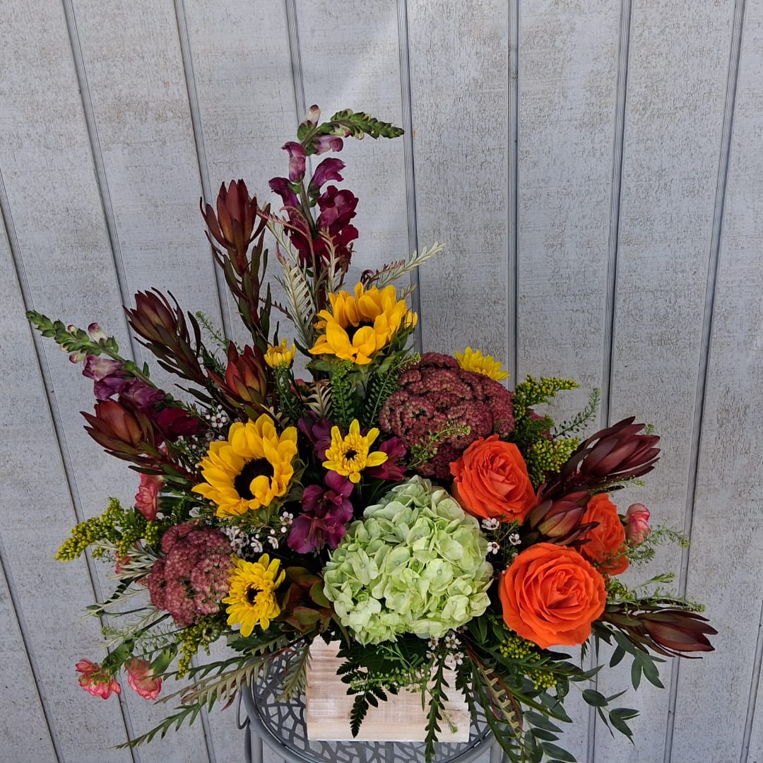 Mixed bouquet with sunflowers, orange roses, hydrangea, and burgundy blooms in a wooden container.
