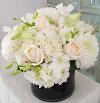 Mona Lisa - A plush cluster of mostly white blooms-big hydrangea heads cushioned by creamy roses, dendrobium orchids and daisy-like blooms-creates a soft, cloudlike bouquet that feels both modern and timeless. This white and pale-green flower arrangement is ideal as an anniversary bouquet or for love and romance, and it also reads as a serene sympathy or wedding arrangement; the rounded, tightly packed shape makes the recipient feel soothed and truly seen.  Set in a black cylindrical vase, the contrast between the luminous white flowers and the sleek black hard good gives the design a clean, contemporary edge that suits birthdays, graduations, thank-you gestures, or simply "just because" moments. The dense layering of hydrangeas, orchids, roses and daisies gives generous visual weight and texture while keeping care simple-keep fresh water in the vase and away from direct heat to help blooms last.