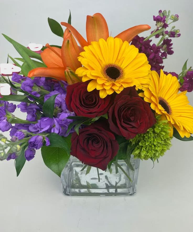Bouquet of yellow gerbera daisies, red roses, orange lilies, and purple flowers in a glass vase