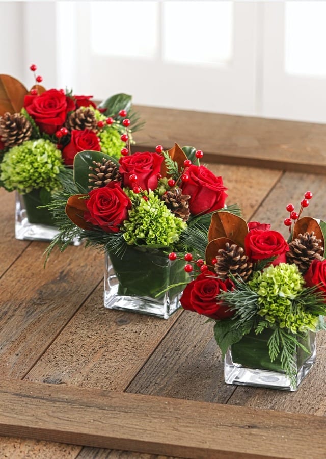 Three red rose arrangements in square glass vases with pinecones and berries