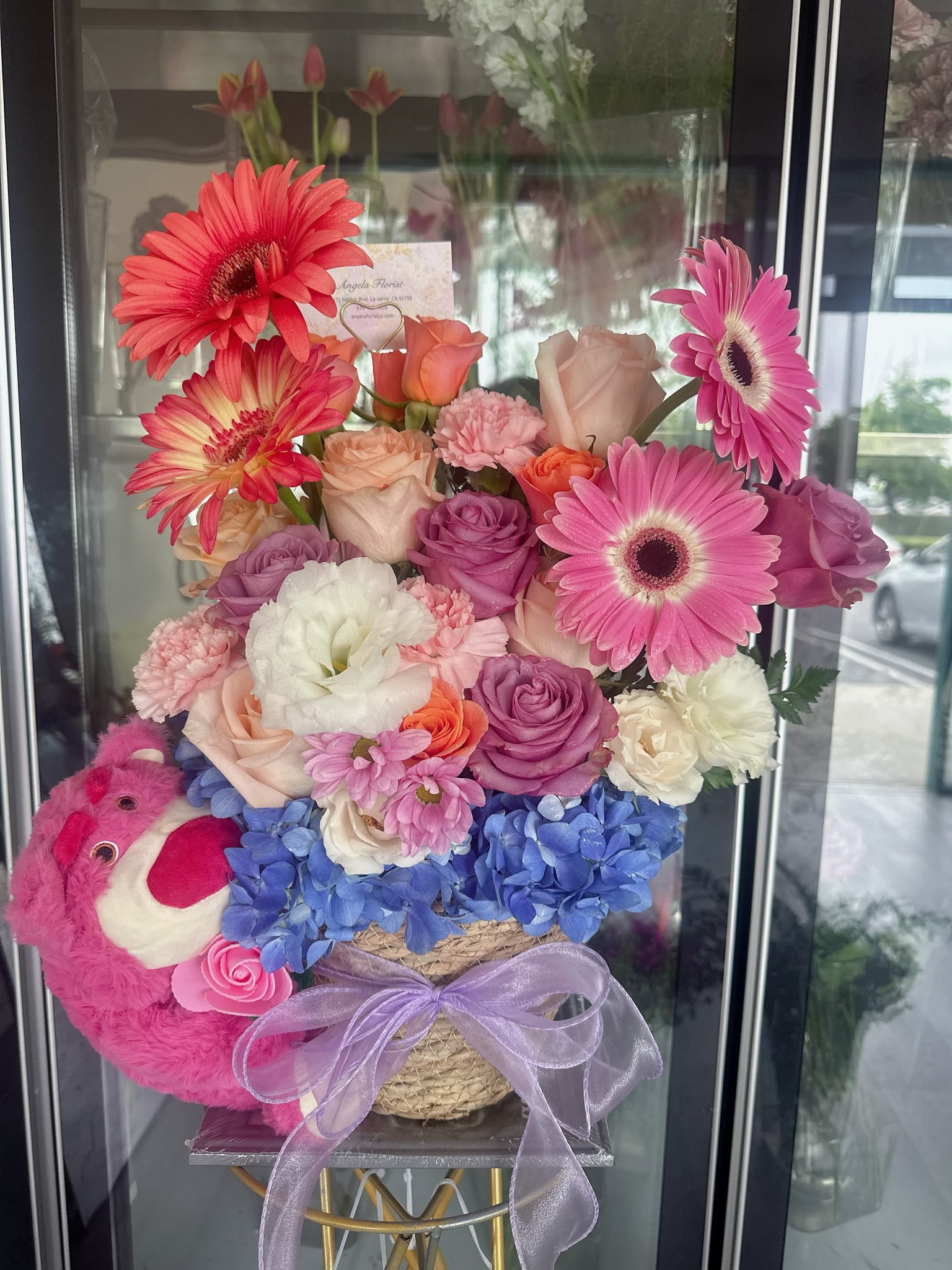 Mixed pink and purple flower arrangement with a plush bear and ribbon