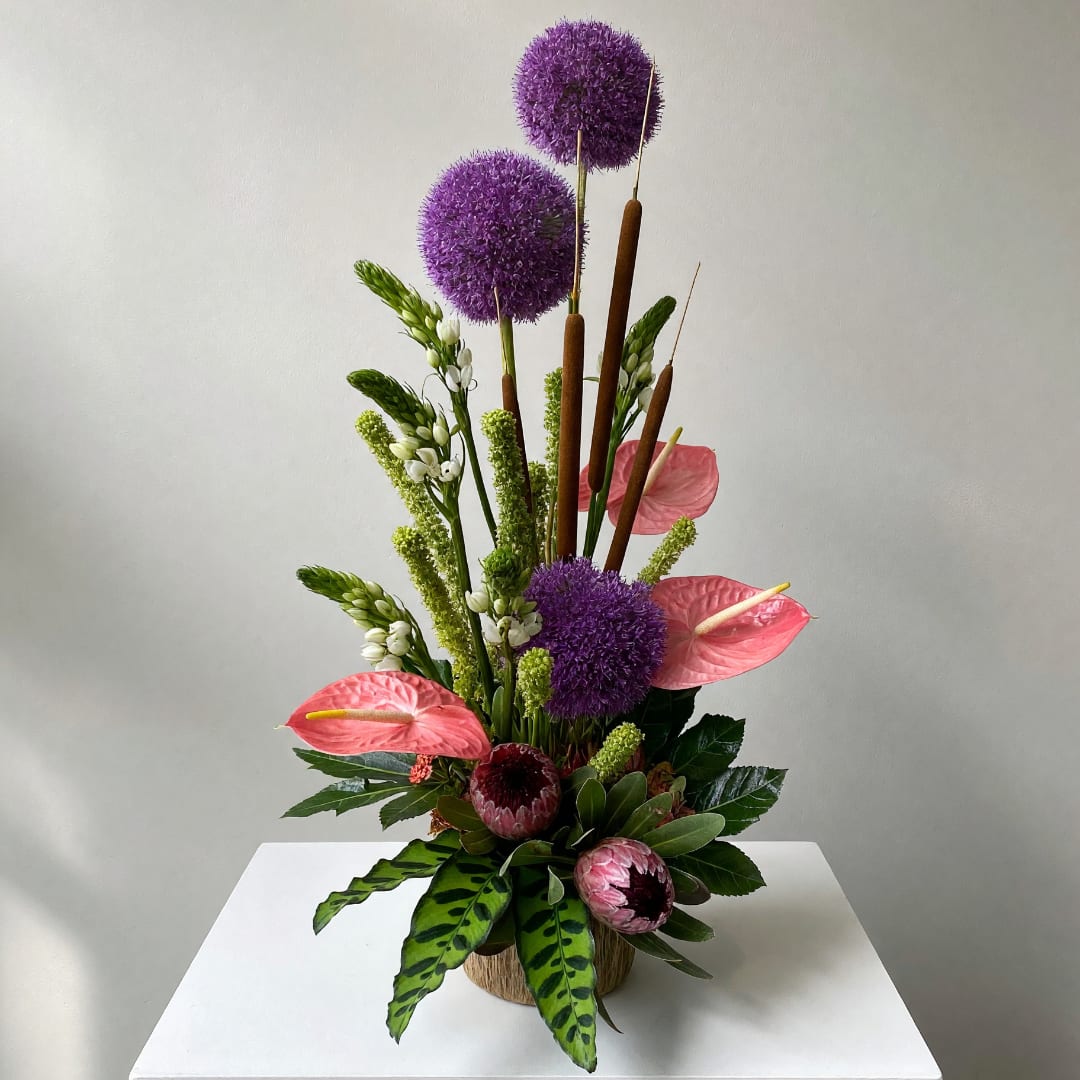 Tall tropical arrangement with purple allium, pink anthuriums, and green accents in a woven container