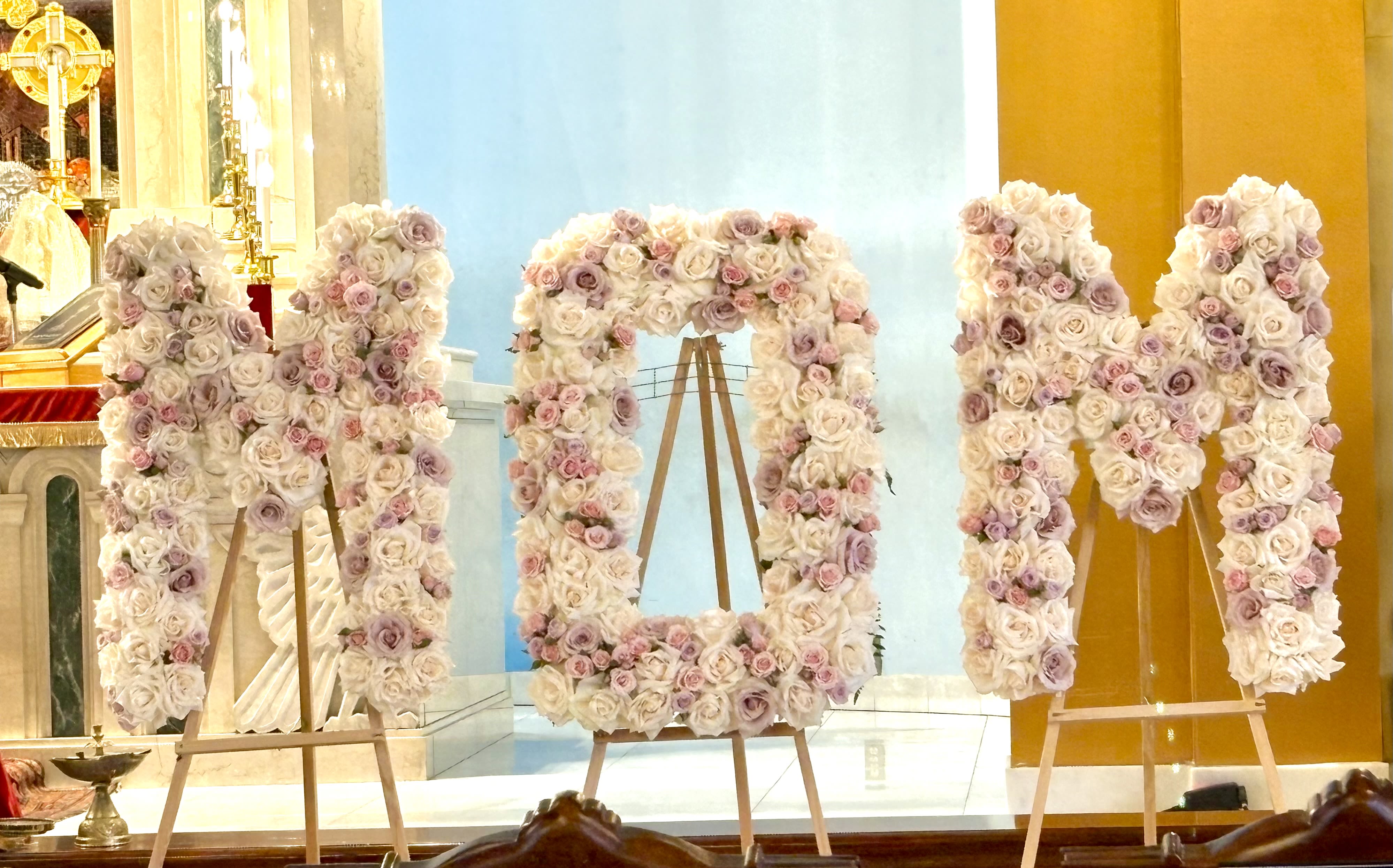 " MOM " By VLM Flowers - Three freestanding letters shaped to spell out a heartfelt tribute are densely covered in pale pink, blush and white roses-an elegant floral display perfect for a ceremony honoring the woman you're celebrating.   Each letter sits upright on its own wooden easel, forming a formal, gallery-style installation that works beautifully as a focal point at a reception, memorial table, or photo backdrop. The roses are tightly packed for a plush, textured surface that reads luxurious from every angle; the overall scale creates a strong presence without overwhelming a space.  Designed to make the person you're honoring feel truly seen, this arrangement suits mothers, grandmothers, and special guests on occasions where a bold, personal statement is wanted. Delivery and placement notes: freestanding letters on wooden easels arrive ready to display; keep in a cool, shaded spot away from direct heat and sunlight to preserve freshness as long as possible.