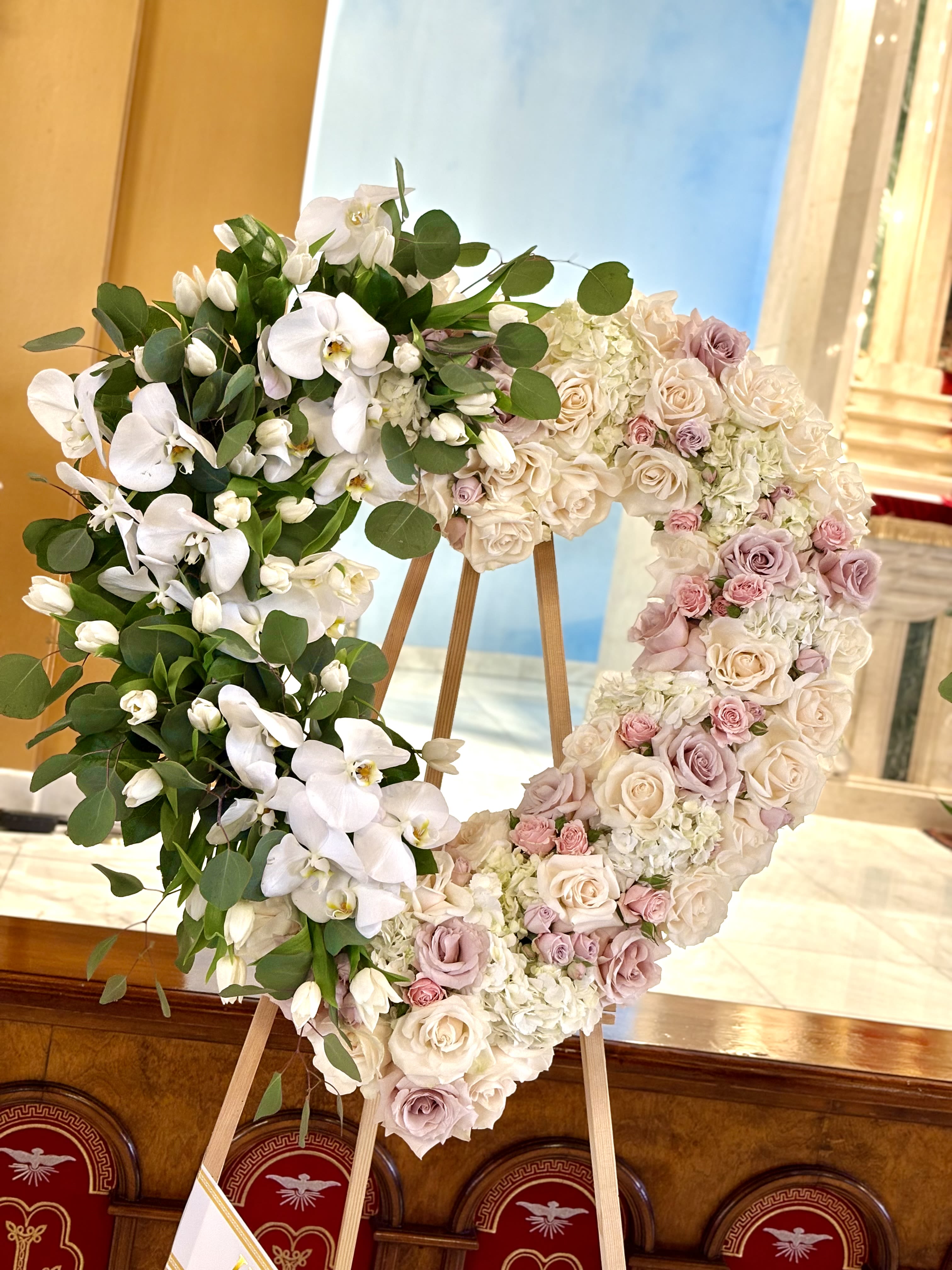 White Orchid Embrace Heart By VLM Flowers - A heart-shaped wreath of soft pastels and white Phalaenopsis orchids creates an immediate sense of gentle grace-perfect as a wedding tribute or a dignified memorial arrangement. This flower arrangement and its formal presentation on a wooden easel send a clear, heartfelt message: the person you're honoring will be remembered with elegance and warmth.  Delicate white orchids sweep down one side of the wreath against deep green foliage, while the remaining surface is richly composed of cream, blush and lavender roses interspersed with pale hydrangea clusters. The mix of textures-silky orchid petals, velvety rose blooms and cloud-like hydrangea-gives the piece visual depth and a soft, romantic palette of white, pink, purple and green.  Designed for display, the wreath arrives ready on a sturdy wooden easel so it can be placed at a ceremony or reception with confidence. The size and heart shape make it an ideal focal tribute for weddings or memorial services, offering the person you're honoring a timeless, soothing expression of love and remembrance.