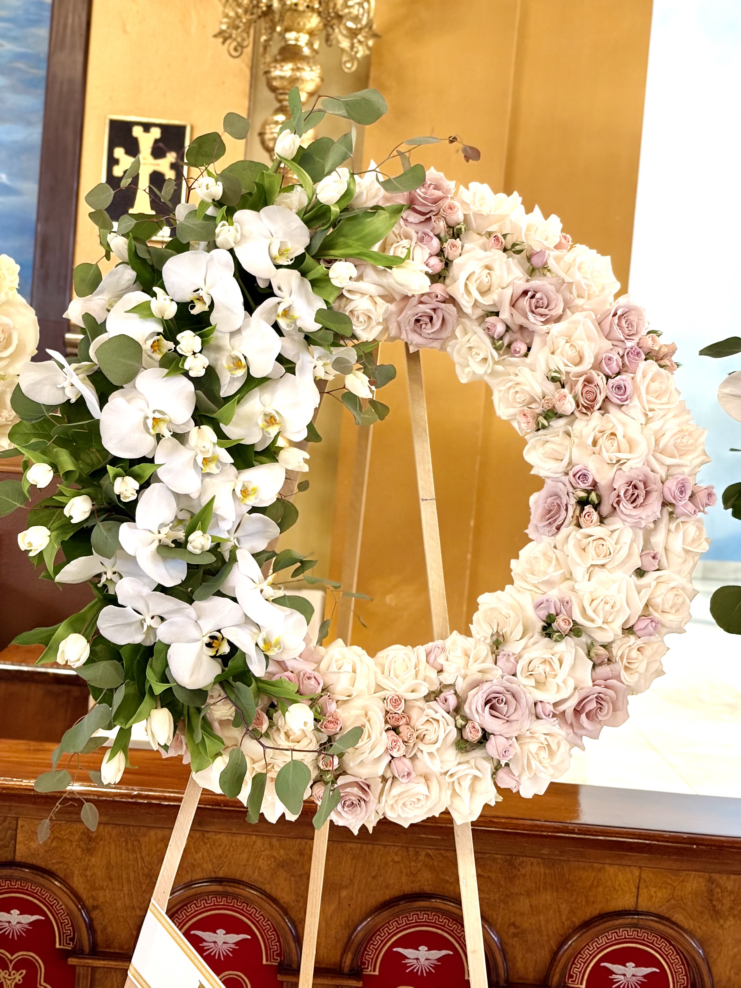 White Orchid Embrace By VLM Flowers - A lush wreath arranged for ceremony and remembrance, this flower arrangement features cascading white Phalaenopsis orchids that spill gracefully down one side-an elegant focal point for a wedding tribute or sympathy display. The white orchid and heart form immediately signal a tender, formal sentiment, making it a meaningful choice for memorial services, funerals, or solemn celebrations of life.  The rest of the heart is densely packed with cream, blush and lavender roses in mixed bloom sizes, accented with eucalyptus and soft green foliage for contrast and texture. Mounted on a sturdy wooden easel, the piece reads large and polished from a distance yet reveals delicate details up close: the glossy orchid petals, the layered rose heads, and the muted pastel palette that feels both romantic and refined.  Designed to honor and comfort the person you're thinking of, this arrangement presents a respectful, heart-centered message at ceremonies or graveside services. Because it's assembled for display, no vase is needed-simply position the easel at the venue; keep orchids and roses away from direct heat and drafts to preserve freshness through the service.