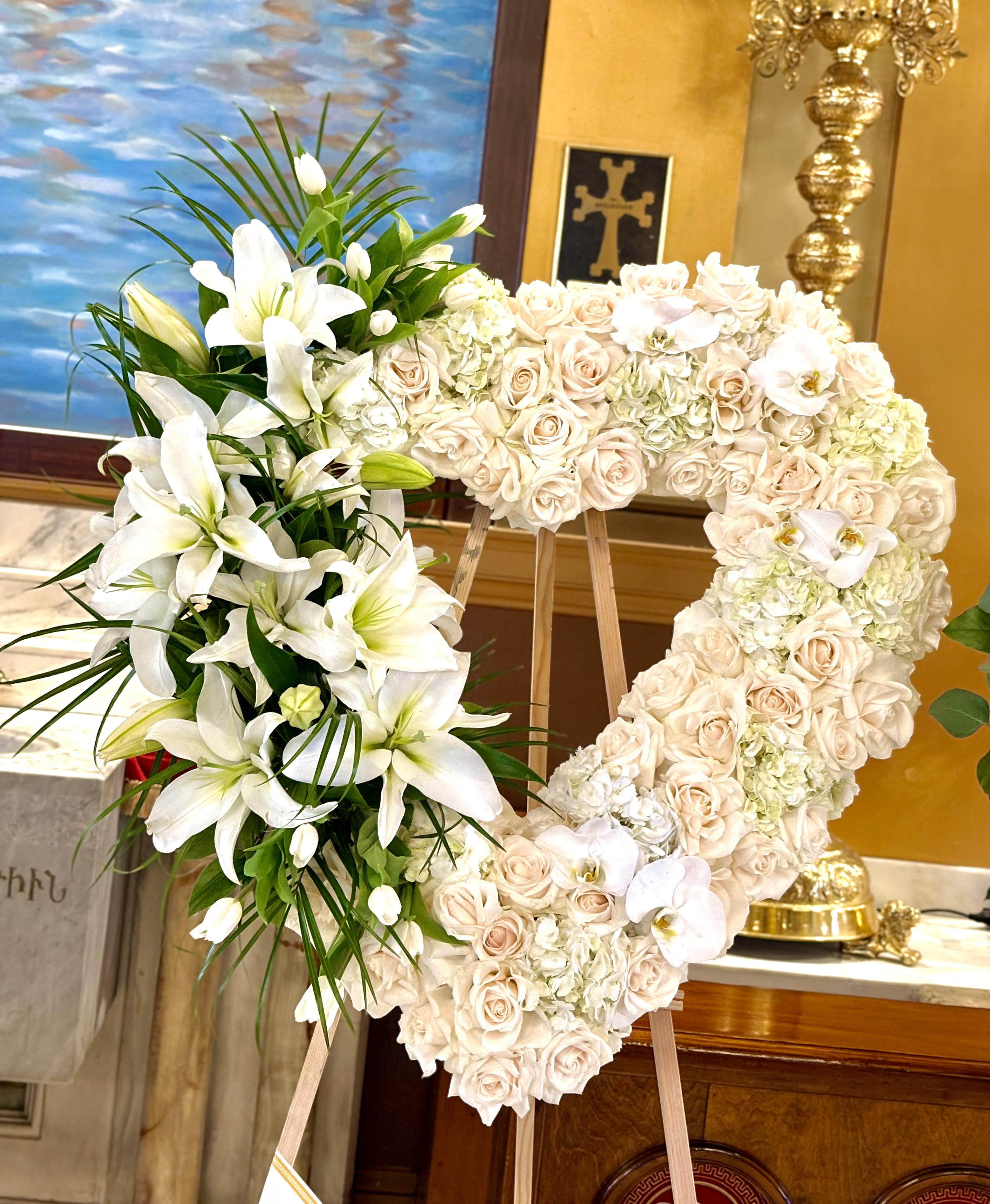 White Heart By VLM Flowers - A large heart-shaped floral wreath that reads as a gentle, timeless tribute-perfect as a sympathy arrangement or a refined wedding memorial. The White Heart by VLM Flowers is arranged on a sturdy wooden easel so the person you're honoring will be remembered with a classic, ceremonial presentation that feels both comforting and elegant.  Clusters of pale blush roses and lush white hydrangeas form the soft foundation, while prominent white lilies and a few white Phalaenopsis orchids provide sculpted focal points and a touch of graceful texture. The white, pink, and green palette creates a peaceful, romantic look that photographs beautifully and reads clearly from a distance.  Made to stand at full height on the included easel, this heart-shaped display is ideal for funeral services, memorial gatherings, or as a wedding tribute that comforts guests. Lightly mist blooms with water before placing indoors; delivered fully assembled for easy placement at the service.