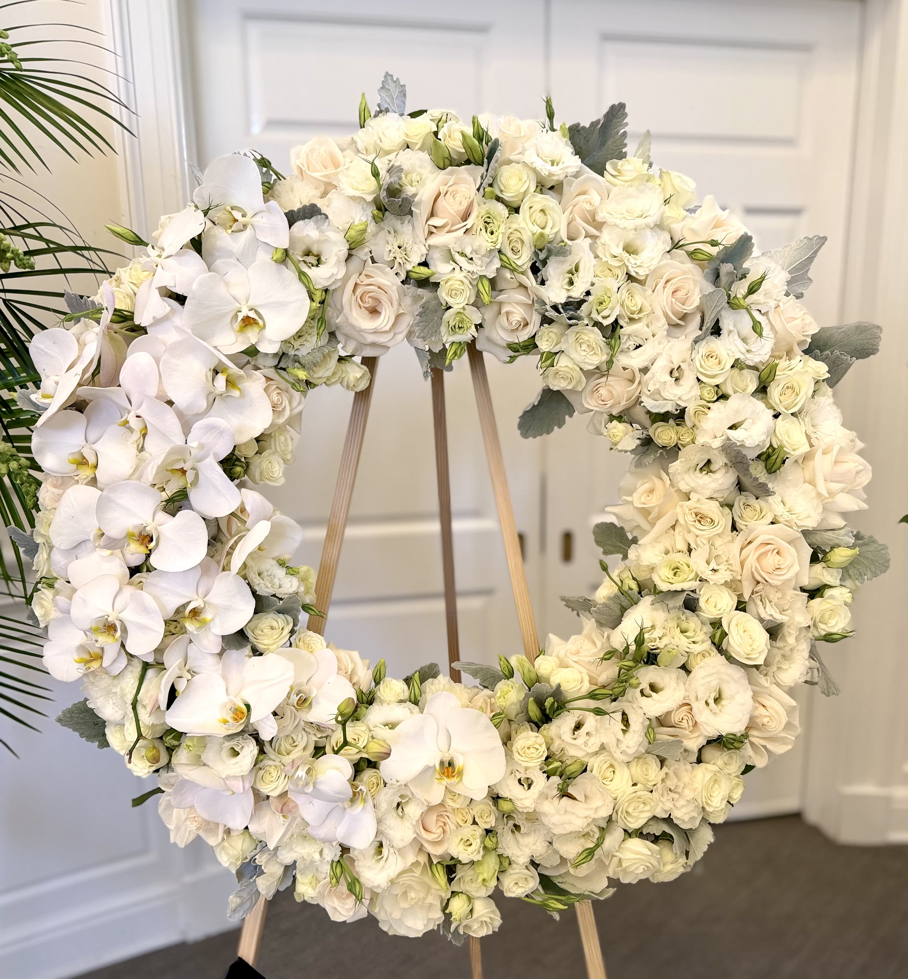 Heavenly Garden Tribute By VLM Flowers - A large circular sympathy wreath arranged with creamy white and soft pink blooms creates a serene, comforting tribute for a memorial service or funeral. This funeral wreath features Phalaenopsis orchids, roses, and lisianthus arranged in a dense ring and presented on a wooden easel stand-an elegant choice when you want the person you're honoring to be remembered with grace and quiet beauty.  Clusters of pure white and cream roses and lisianthus are interspersed with delicate blush roses and dramatic white Phalaenopsis orchids, while silvery foliage adds muted contrast and soft texture. The wreath's full, packed composition reads formal and refined from every angle; its large circular shape provides a classic, reassuring silhouette suitable for chapel, graveside, or reception display.  Choose this arrangement to convey sympathy, respect, or lasting remembrance-it's the kind of tribute that helps mourners feel held and seen. The wreath arrives ready to stand on the included wooden easel; minimal care is needed beyond keeping it cool and sheltered until the service.