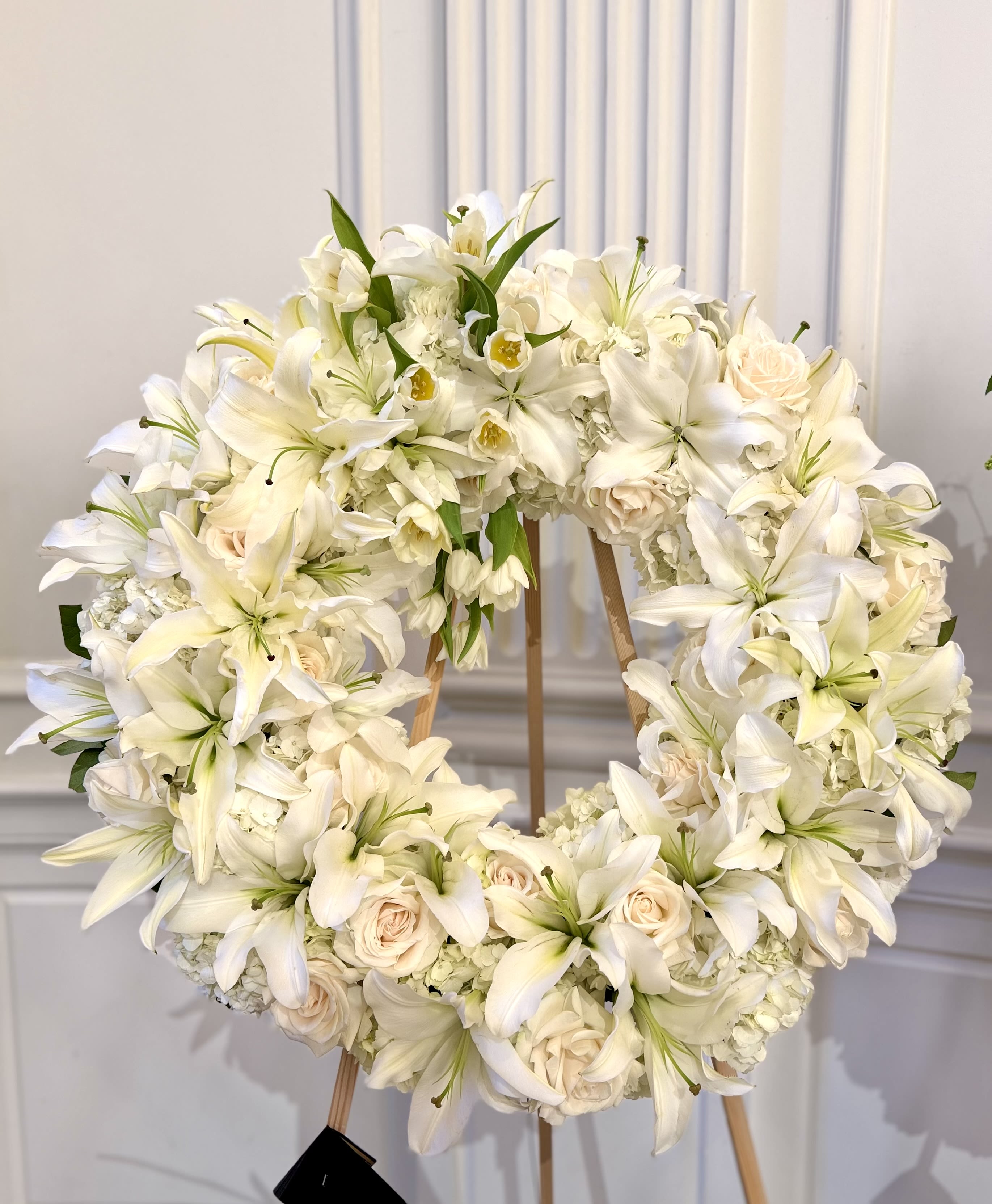 Eternal Grace Wreath - A large circular sympathy wreath arranged with serene white lilies, pale blush roses, and soft clusters of white hydrangeas-designed to convey calm and comfort at a funeral or memorial service. This flower arrangement for sympathy and remembrance sits upright on a wreath stand, presenting a formal, graceful tribute that helps the person you're honoring feel quietly celebrated.  The design is full and layered, with flowers packed closely together to create a plush, continuous surface in white, pale pink, and fresh green foliage. Lilies provide sculptural height and a gentle fragrance, roses add tender blush tones, and hydrangea clusters bring soft texture across the wreath's broad face. Sized for display rather than carrying, it arrives ready to place on the included stand for immediate presentation.  Appropriate for funerals, memorials, and services of remembrance, this wreath expresses sympathy without words-the kind of arrangement that offers solace to family and friends. The stand is included for ease of setup; keep the flowers in a cool location away from direct sun until the service to preserve their form and freshness.
