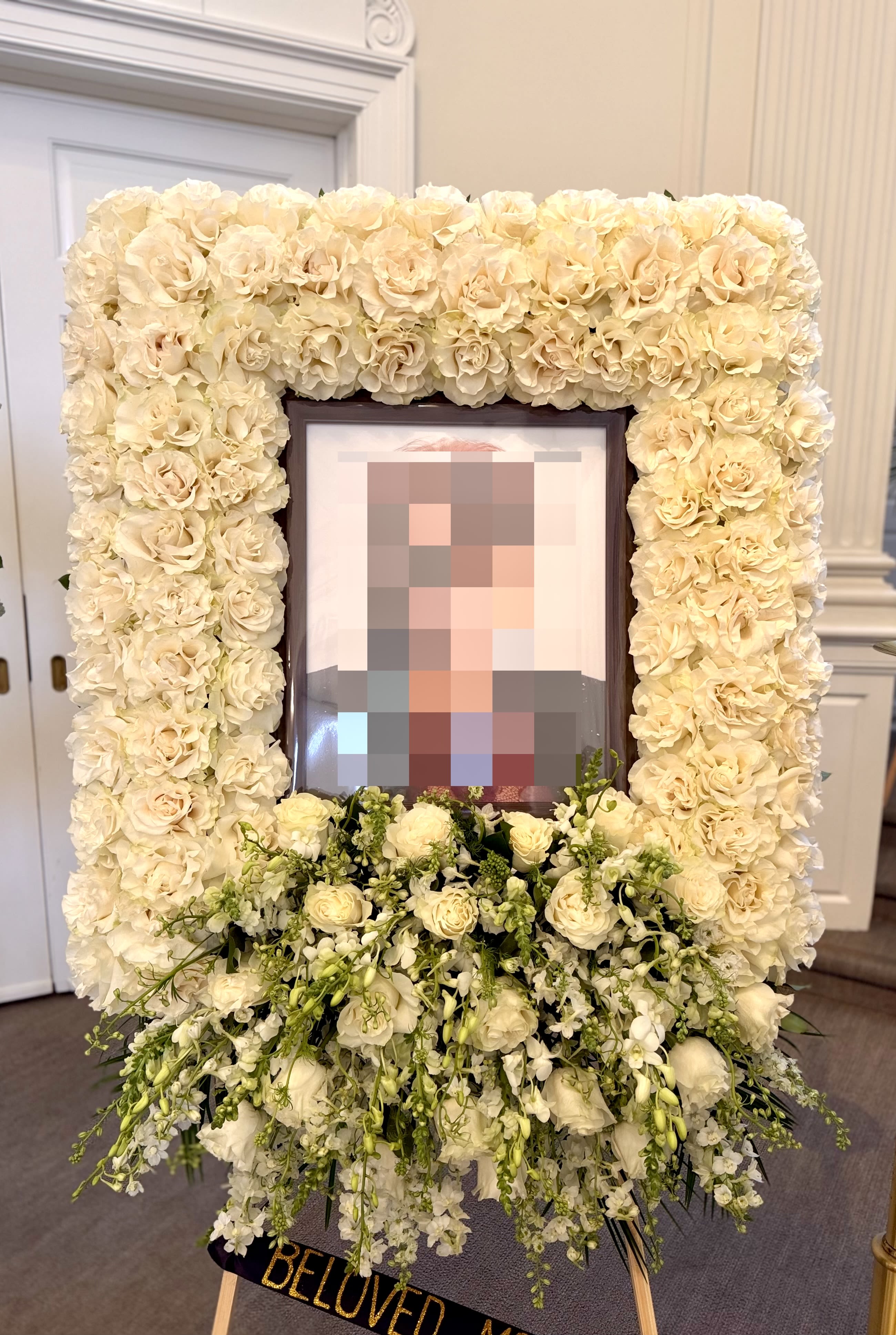 A Lasting Embrace By VLM Flowers - A densely packed rectangular frame of white roses surrounds a treasured portrait, creating a quietly powerful tribute arrangement for sympathy and funeral services. The white-and-green palette reads serene and respectful, the formal composition giving the person you're honoring a sense of dignity and calm. This is the kind of memorial display that helps mourners focus on memories and comfort.  Beneath the rose-bordered frame a graceful cascade of white blooms and trailing green accents softens the base, adding depth and gentle movement to the piece. Orchids and snapdragons are woven into the design alongside other complementary flowers for texture and visual interest, while a tasteful ribbon banner and easel complete the presentation. A small picture frame holds the portrait securely at the center so the face of the loved one is the focal point.  Made to stand at services or beside a memorial table, this arrangement communicates sympathy and reverence-the kind of tribute that lets family and friends feel seen and supported. Care and sizing details are provided at checkout; our team will handle delivery and placement so the arrangement arrives fresh and respectfully prepared for the service.
