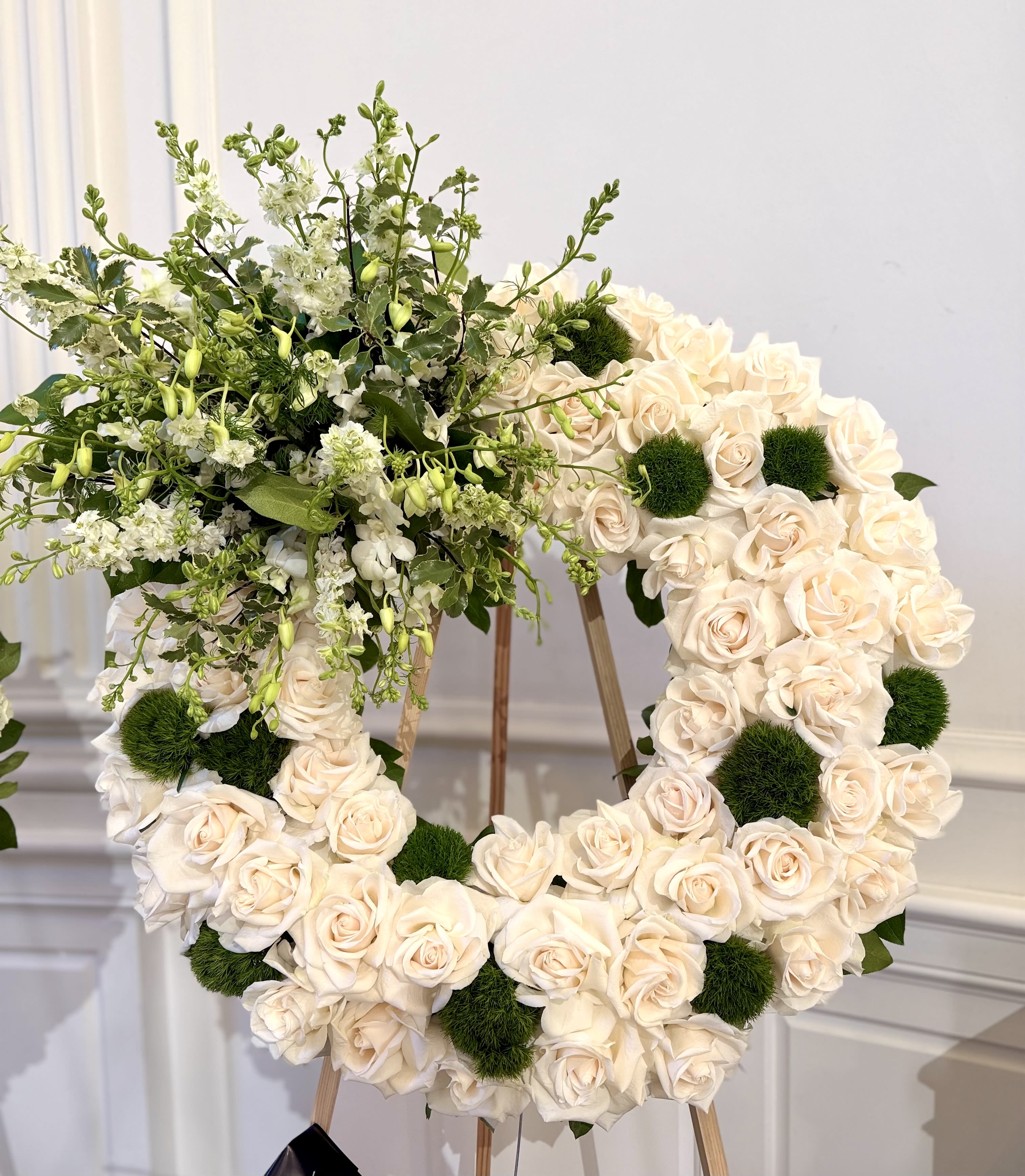 Blush Garden of Grace By VLM Flowers - A large wreath of cream  roses creates an immediate sense of tender remembrance - perfect as a sympathy memorial piece or a formal tribute for a funeral. The form and soft color palette say "you are in our hearts" in a gentle, dignified way, making the recipient feel honored and comforted in a difficult moment.  This ceremonial arrangement sits on a wooden easel and is composed mainly of cream roses accented with small white filler blooms and airy green foliage concentrated toward the upper left, giving a natural, asymmetrical flourish. Dark green mossy pom-pom details are scattered through the design to add texture and depth against the pale roses; overall the wreath reads soft, romantic, and thoughtfully crafted at a scale meant for display rather than hand-carrying.  For a family service or to mark a memorial, this heart wreath offers a classic, heartfelt expression of sympathy that will help those grieving feel seen and remembered. The easel-mounted format makes delivery and presentation simple; for care, keep the arrangement in a cool, sheltered place until the service and let us know any placement needs so we can ensure it arrives ready for display.
