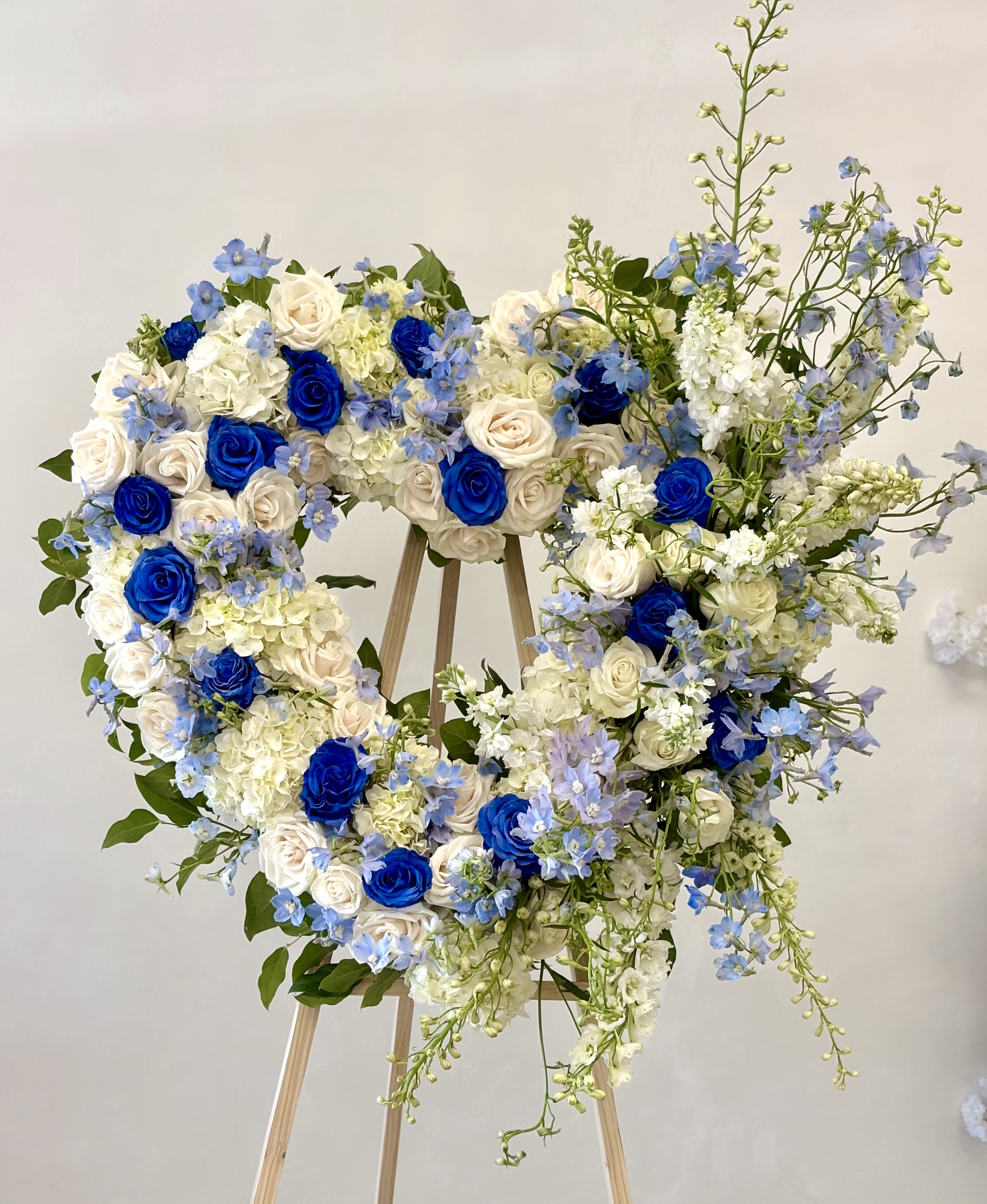 Royal Blue Heart By VLM Flowers - A large heart-shaped wreath in cool blues and whites creates an immediate feeling of calm and reverence - perfect as a sympathy arrangement. The heart silhouette and formal presentation make the recipient feel truly honored; this is the kind of arrangement that quietly says, "we remember" or "you are deeply loved."  Tightly clustered blue and white roses form the core of the design, accented with pale hydrangea heads for soft volume and airy blue delphinium for vertical movement and texture. The composition is full and layered, with taller stems rising on one side to give graceful height, and fresh green foliage woven throughout to brighten the cool palette. Mounted on a sturdy wooden easel stand, the wreath is ready for display at a service, reception, or memorial.  Choose this arrangement when you want the person you're honoring to feel seen and respected - whether marking a solemn moment or celebrating enduring love. Careful construction and live blooms mean the tribute arrives ready to present; please follow standard floral care for cut flowers and contact us with any display questions when ordering.