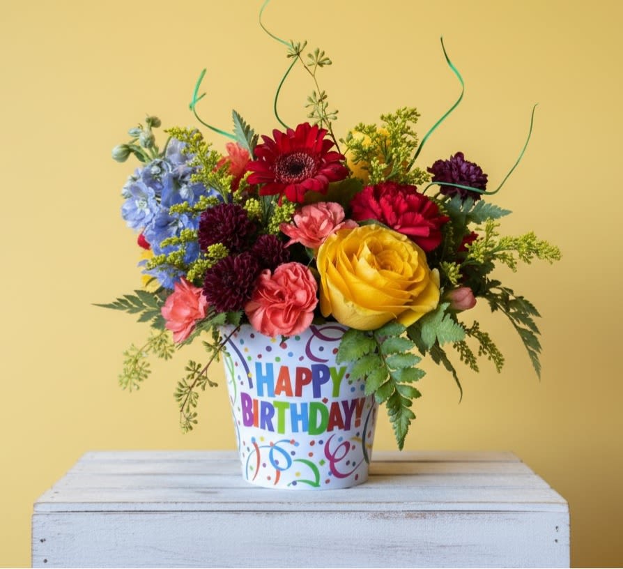 Birthday Blossoms -  The arrangement features a diverse mix of flowers, each contributing to a lively and colorful display. What a way to celebrate ! Designed in a brightly colored melamine container with the words Happy Birthday approx dimensions 10 in X 10 in