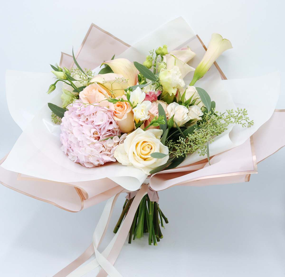 Hand-tied bouquet of cream roses, pink hydrangea, and pale calla lilies wrapped in blush paper.