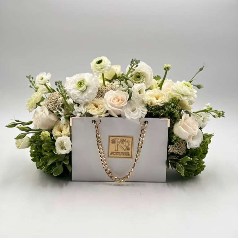 Luxe Garden Handbag - A timeless expression of elegance, this arrangement features a harmonious blend of ivory, soft cream and white blooms, beautifully arranged in a signature handbag-style box with gold chain accents. Lush greenery adds depth and freshness, while the refined neutral palette creates a sense of calm sophistication. Perfect for honoring someone special, this design is a graceful and luxurious way to celebrate love, appreciation, and timeless beauty.