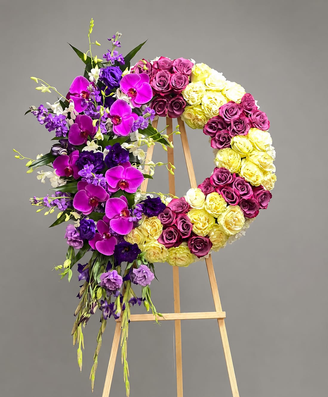 Sympathy Orchical Serenity - This is a graceful sympathy wreath designed to honor a life beautifully lived. Featuring luxurious lavender and ivory roses, vibrant purple orchids, elegant lisianthus, and cascading seasonal blooms, this handcrafted tribute symbolizes eternal love, peace, and cherished memories. The circular design represents the everlasting bond between loved ones, while the flowing floral accents add a sense of serenity and hope. Thoughtfully created by Kenneth Village Flowers, this elegant arrangement offers a heartfelt expression of sympathy, comfort, and remembrance during life's most difficult moments.  A printed ribbon or memorial card is included. Please indicate your preference in the “Special Instructions” section at checkout. They can be customized with your message that you can write in the "Card Message" section at checkout. Memorial cards are recommended for longer messages and personal notes while ribbons are best suited for short wording such as senders' names.