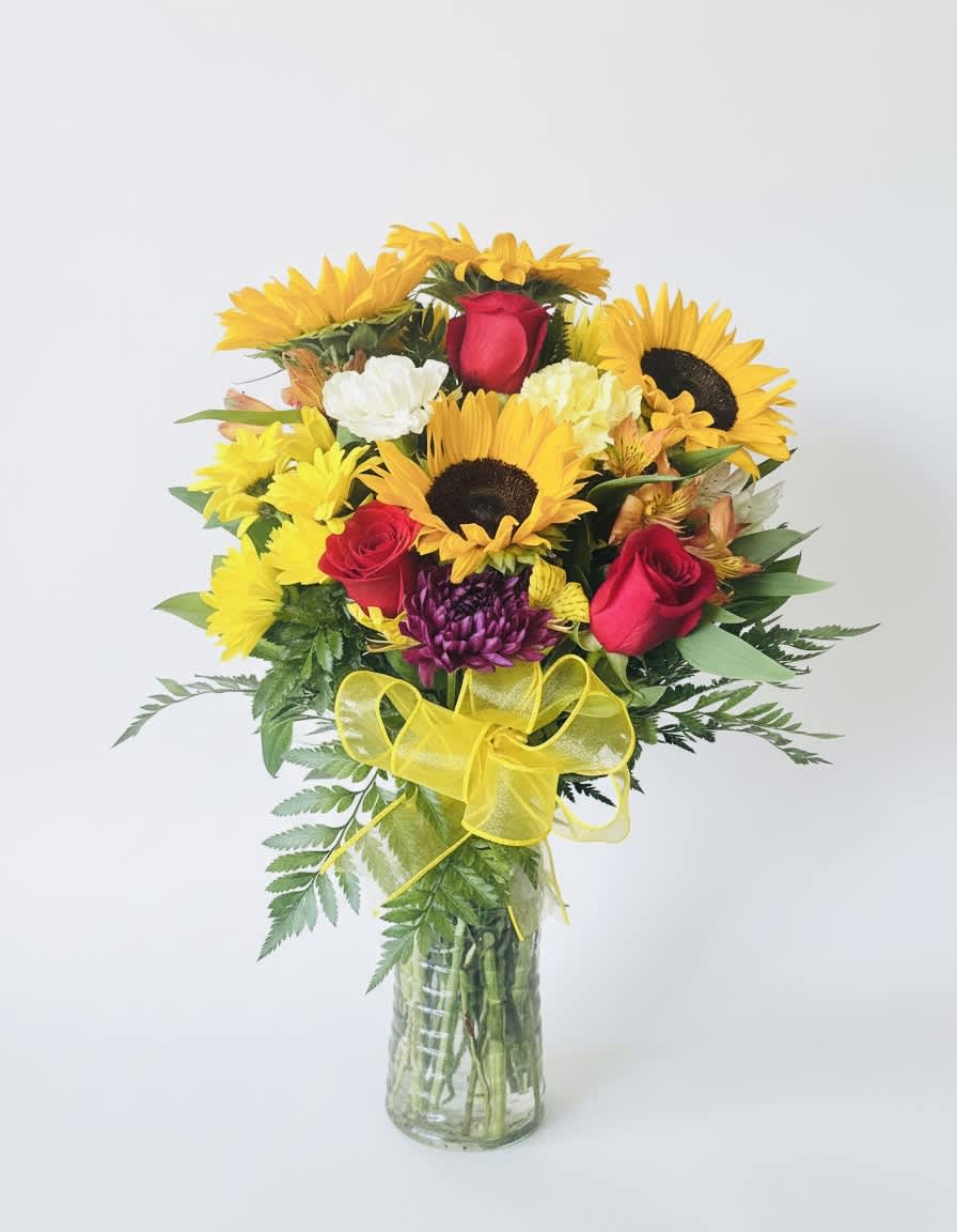Bouquet of sunflowers, red roses, and mixed blooms in a glass vase with a yellow bow