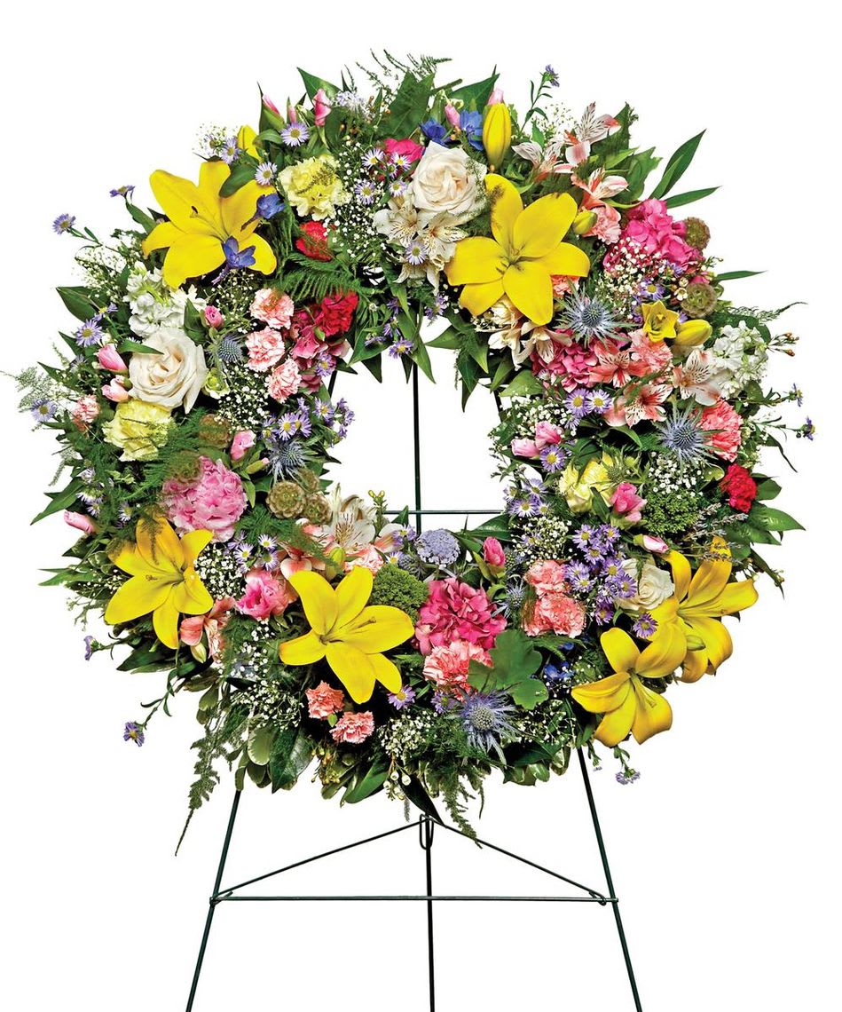 Warm Thoughts Wreath - A bright standing wreath of greenery and fresh flowers in yellow, pink, blue and white will send a message of hope and harmony, and help to celebrate a meaningful life. Standard size 18" diameter form used. Deluxe size 24" diameter form used.