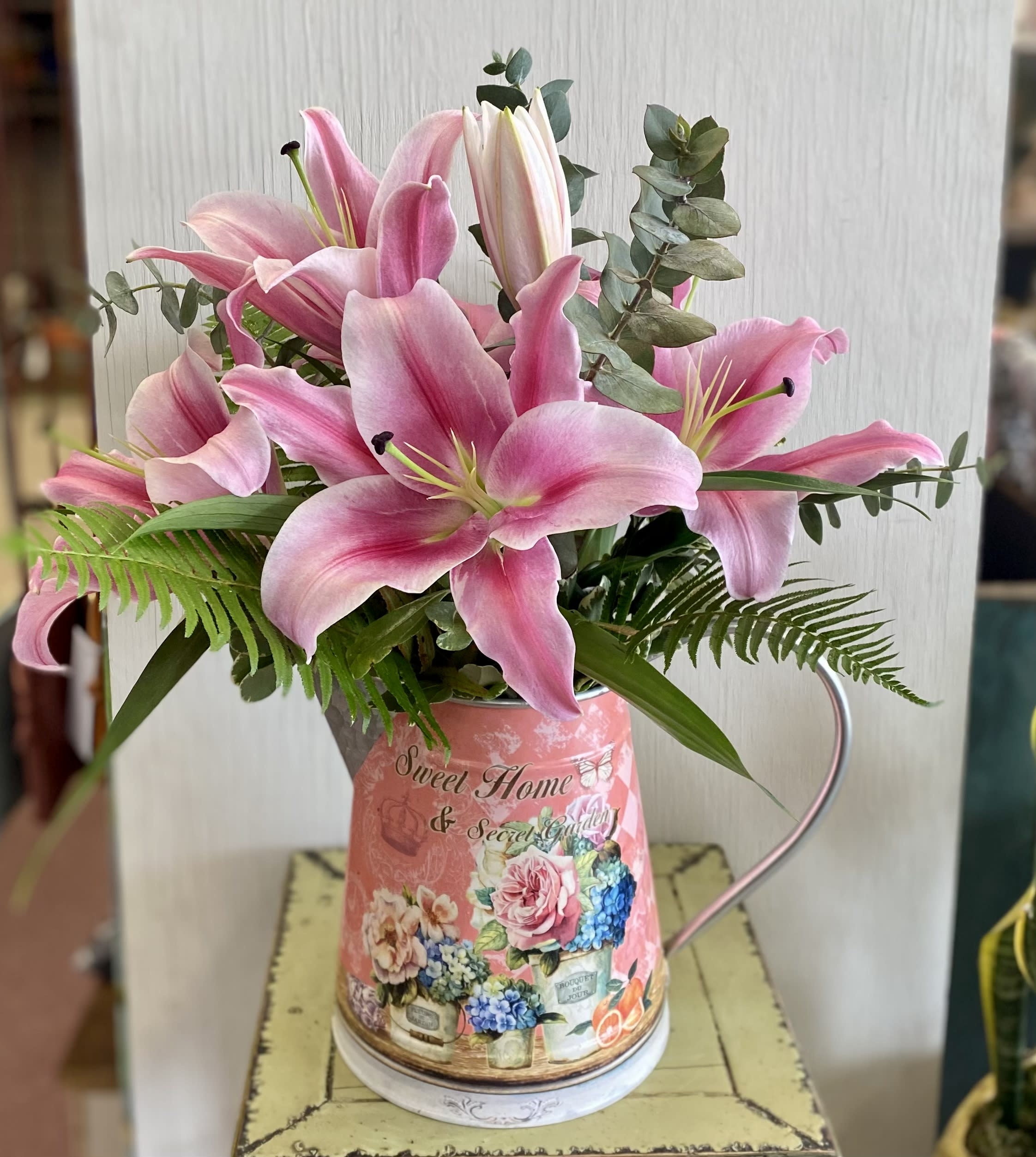 Pink lilies arranged in a decorative metal pitcher with greenery