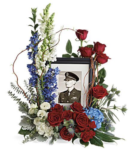Floral tribute around a framed military portrait with red roses and blue-white flowers