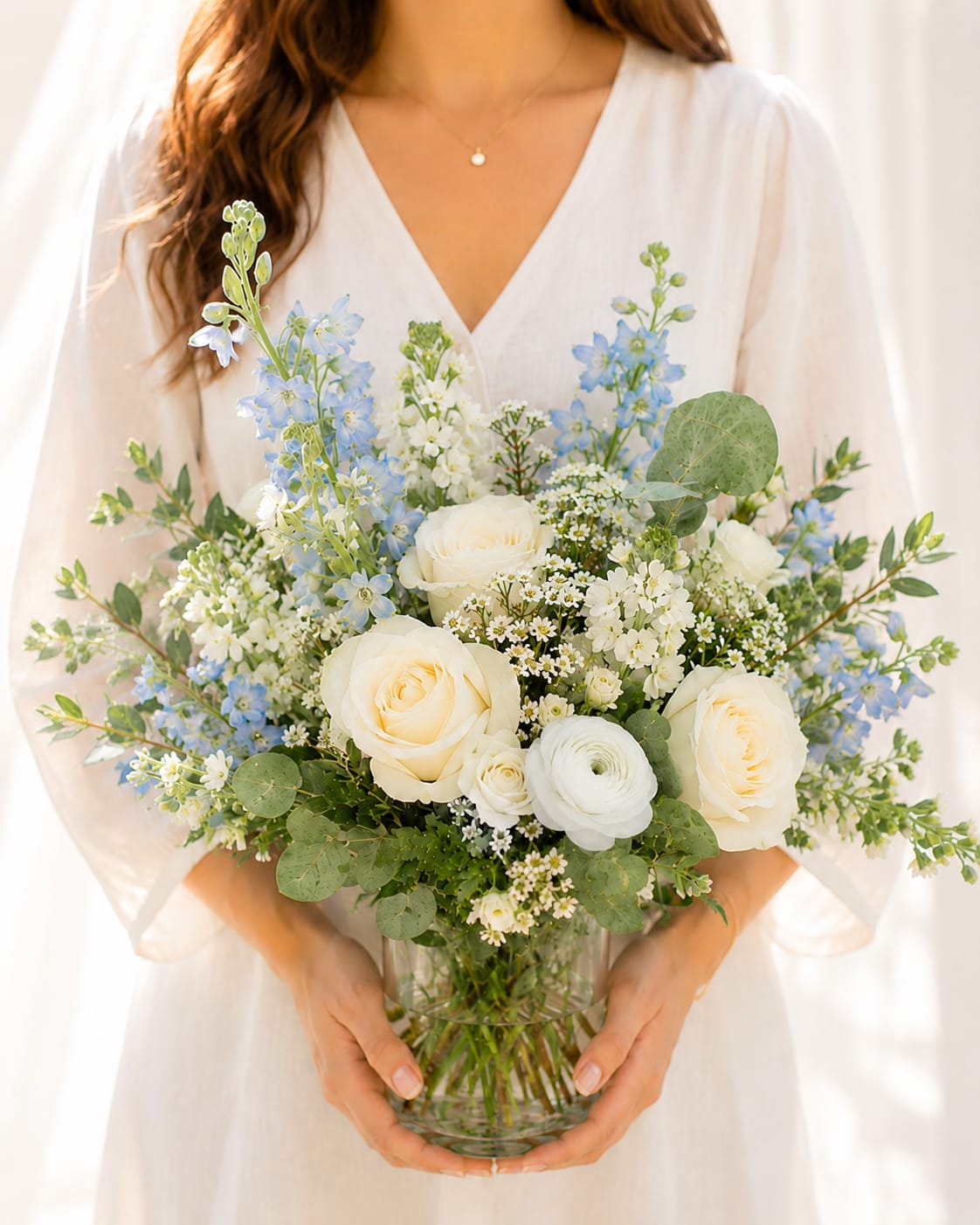 Petite White &amp; Blue Garden Arrangement - 12” tall. Creamy white roses and airy blue delphinium-like. The pale palette reads fresh and elegant, and the loose garden-style shape feels effortless-the kind of arrangement that makes the person you're celebrating look and feel luminous on a special day.  eucalyptus leaves and small white filler flowers add muted green contrast and delicate texture, arranged in a clear glass vase that lets the blooms float naturally. The composition is light and airy with a gentle, rounded silhouette that suits wedding tables, a bridal suite, or a meaningful at-home gift.  Ideal for anyone you want to honor on an important occasion, this bouquet sends a message of serene beauty and calm joy. Vases arrive ready to display; change the water every couple of days and trim stems at an angle to keep the roses and delphinium looking fresh longer.