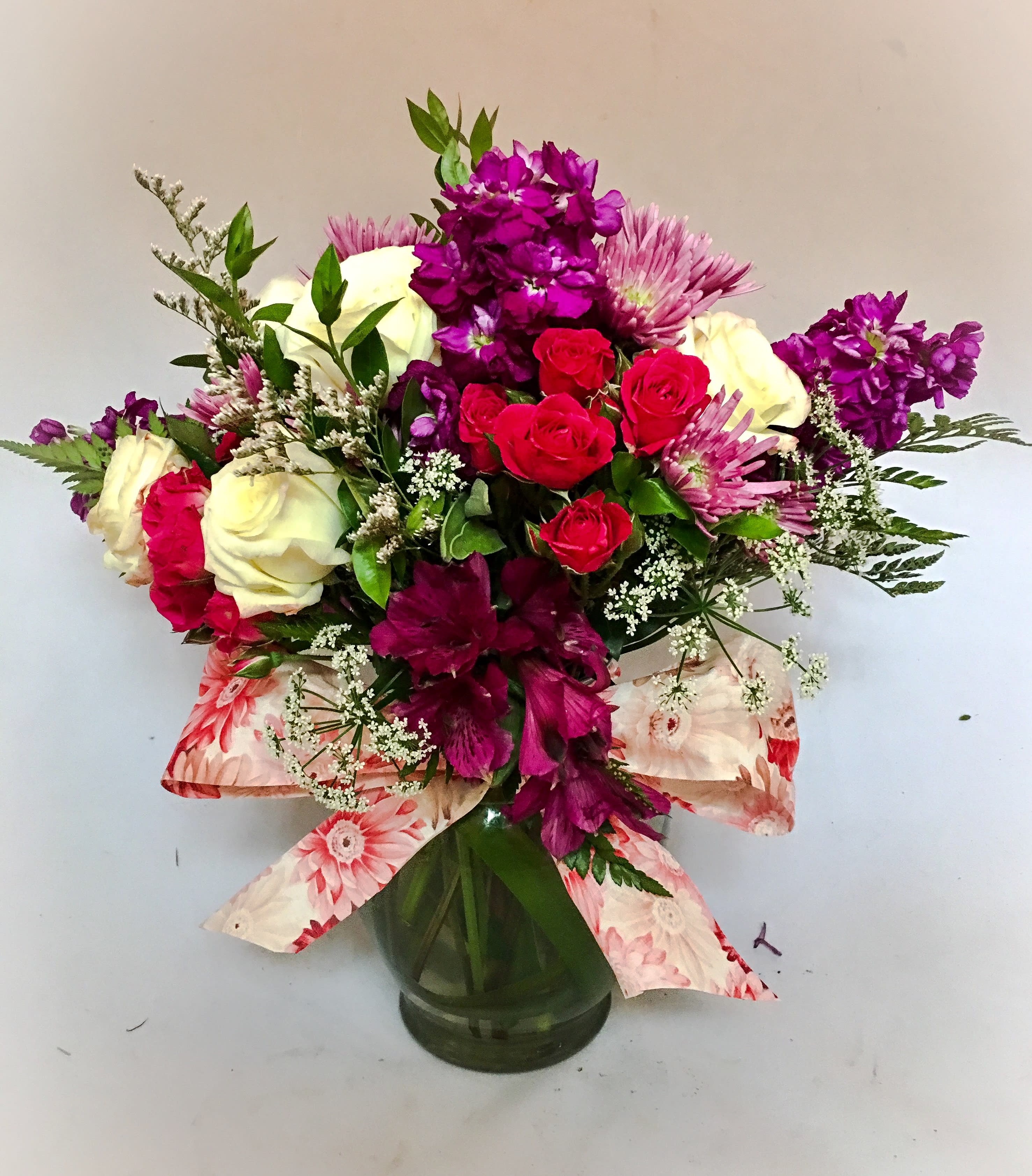 Mixed arrangement of white roses, red spray roses, and purple flowers in a glass vase with a floral ribbon bow