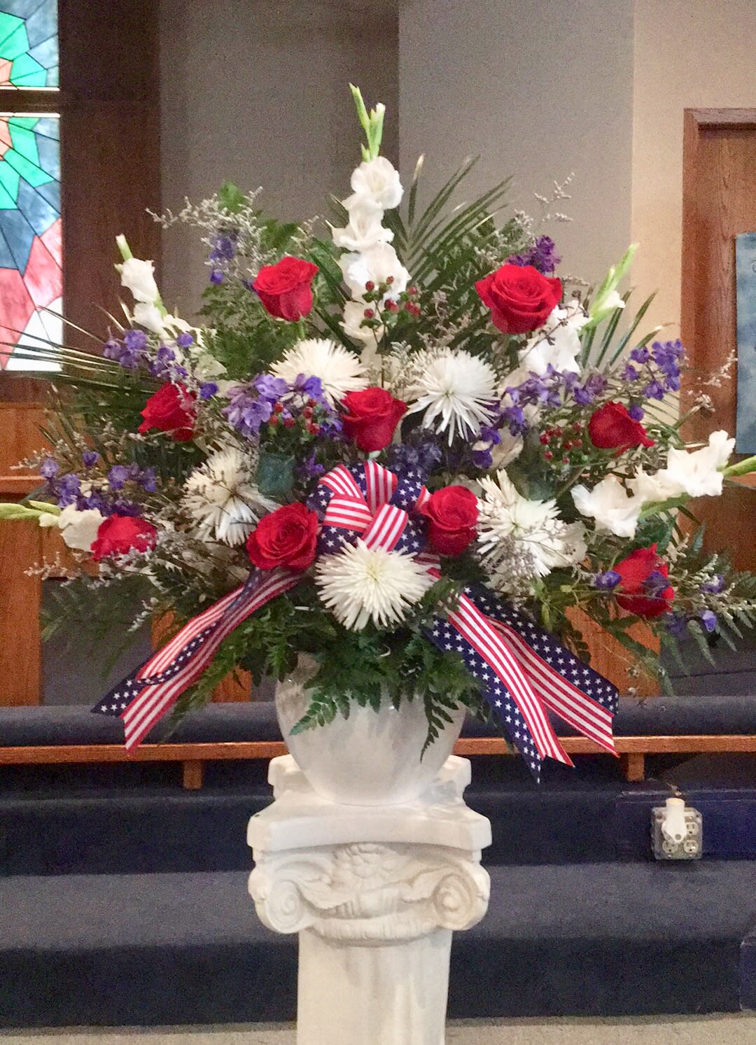 Tall red, white, and purple flower arrangement in a white urn with patriotic ribbon on a pedestal