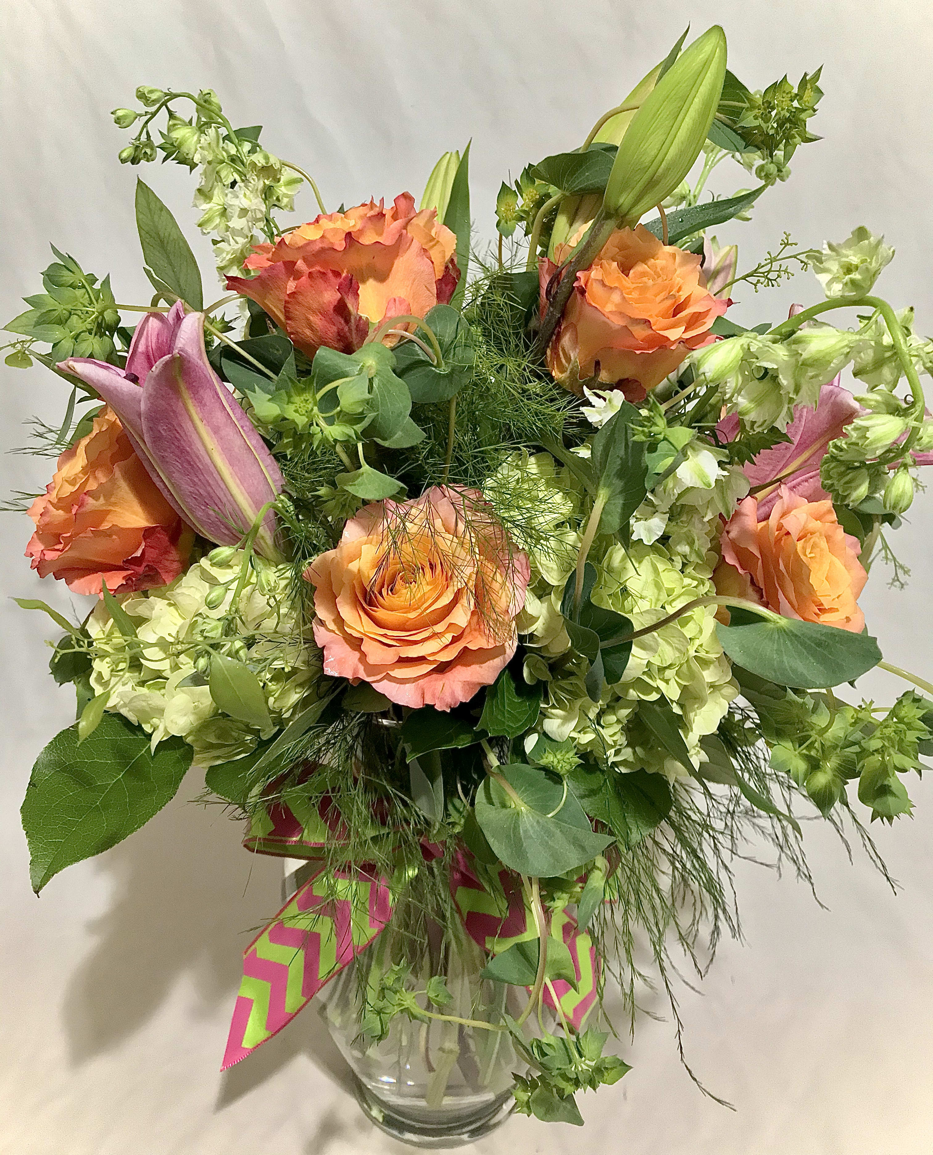 Tall arrangement of orange roses, pink lilies, and green hydrangeas in a clear glass vase