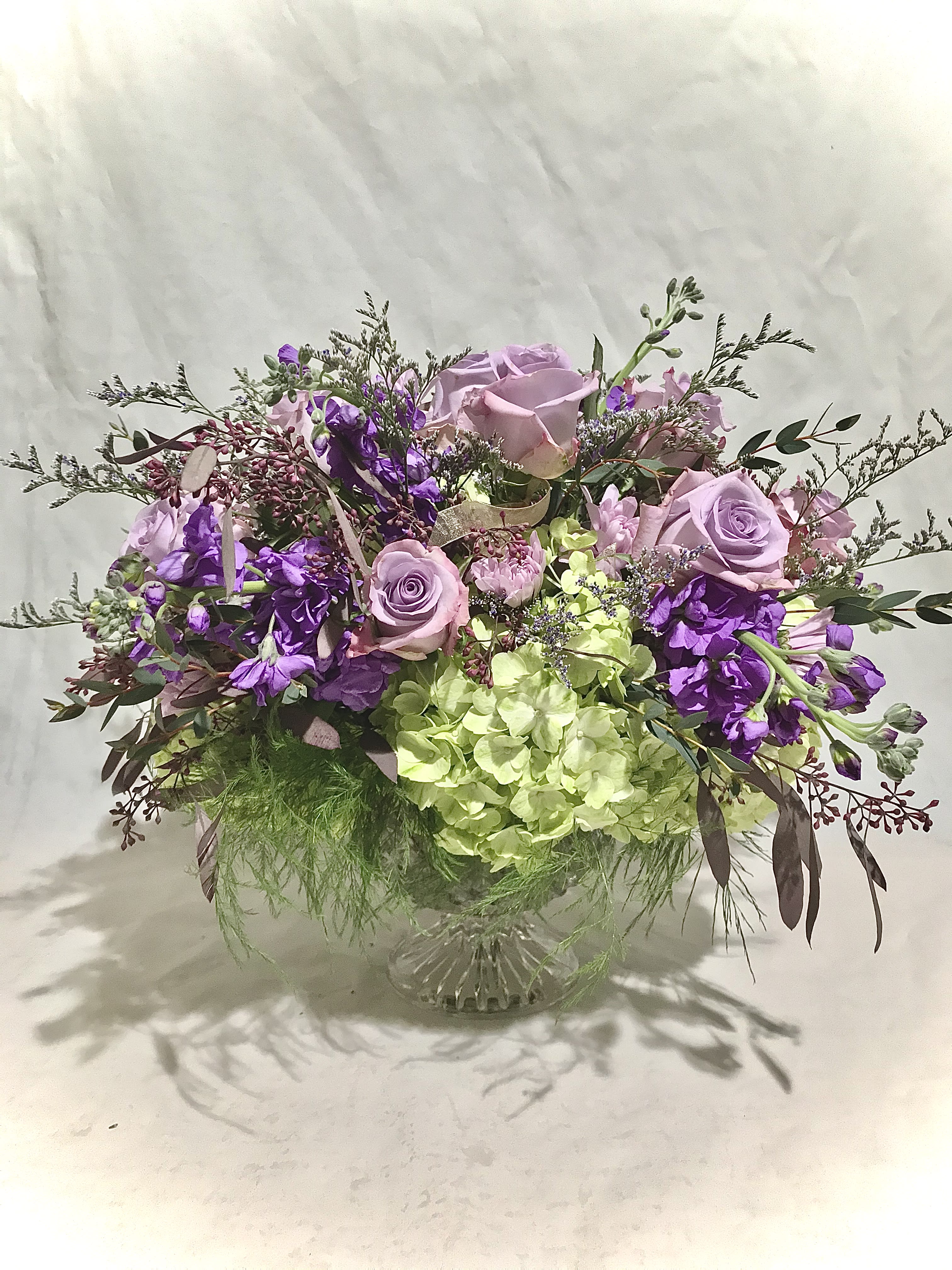 Low pedestal arrangement of lavender roses, purple blooms, and green hydrangeas in a glass compote vase