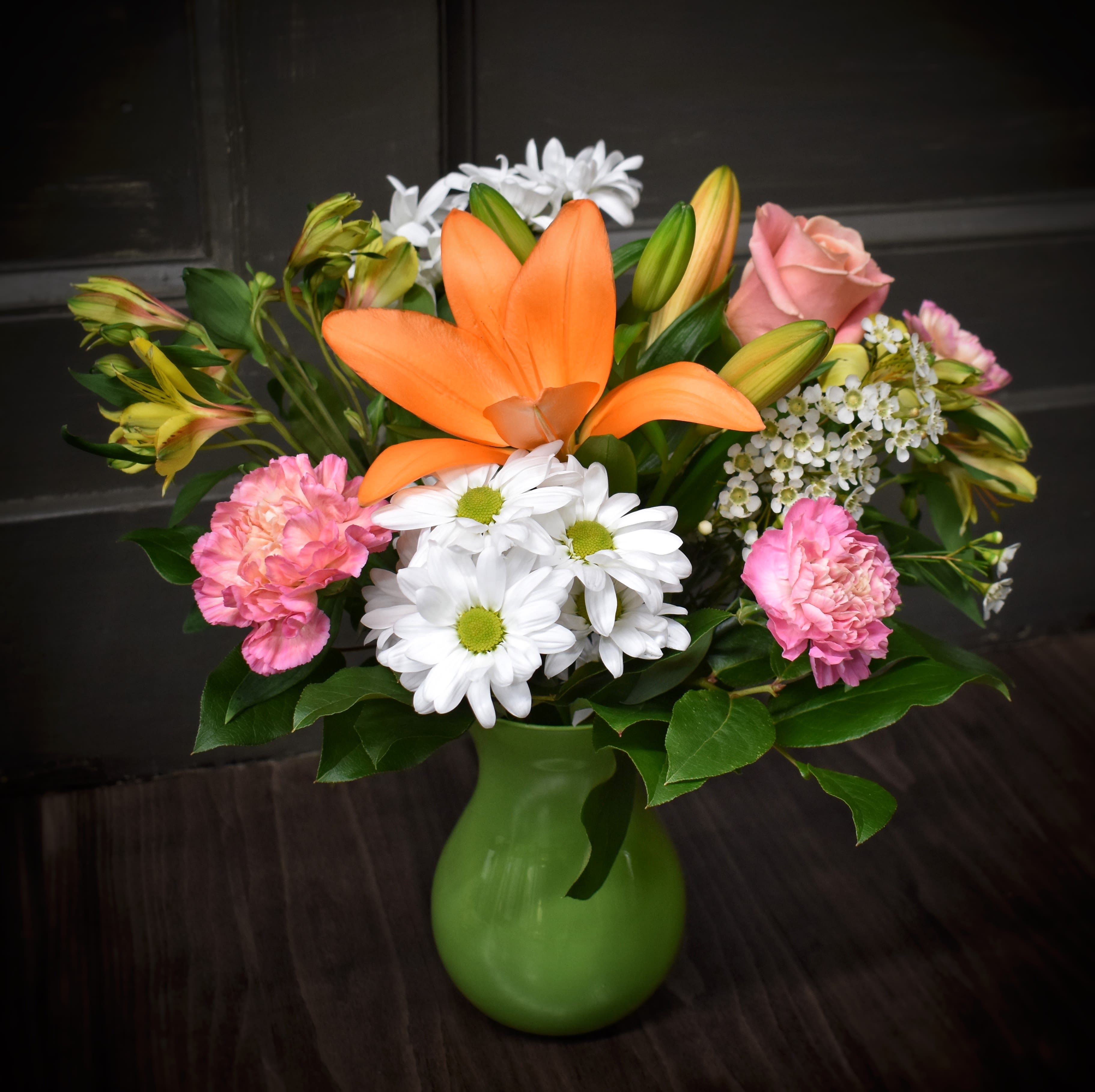 Designer's Choice Traditional - Bright, garden-fresh color greets the recipient the moment they see this Designer's Choice Traditional flower arrangement - a cheerful bouquet perfect for birthdays, everyday gifting, or a heartfelt thank-you. Whether the focal orange lily and pink rose in a bold green ceramic vase or the bright orange gerbera and soft pink roses in a clear glass vase, the mix of orange, pink, white, green and purple reads as joyful and uplifting, the kind of arrangement that makes someone feel genuinely seen and celebrated.  Carefully mixed blooms include lilies, roses, carnations, alstroemeria, daisies, and gerbera daisies with accents of eucalyptus and leafy greens for texture and softness. One presentation arrives in a rounded green ceramic vase for a vivid, modern contrast; another is styled in a clear glass vase showcasing stems and a medium, hand-arranged fullness. Both options create layered shapes - rounded blooms and spiky buds - that give visual energy and a fresh garden scent.  Ideal for sending to a friend, colleague, or loved one who deserves a bright pick-me-up, this design balances playful color with romantic touches so it fits many occasions from celebrations to simple thinking-of-you moments. Arrangements are hand-designed by our florists; vase choice reflects availability and the pictured styles, and standard care is to keep flowers in fresh water and trim stems every few days to prolong enjoyment.