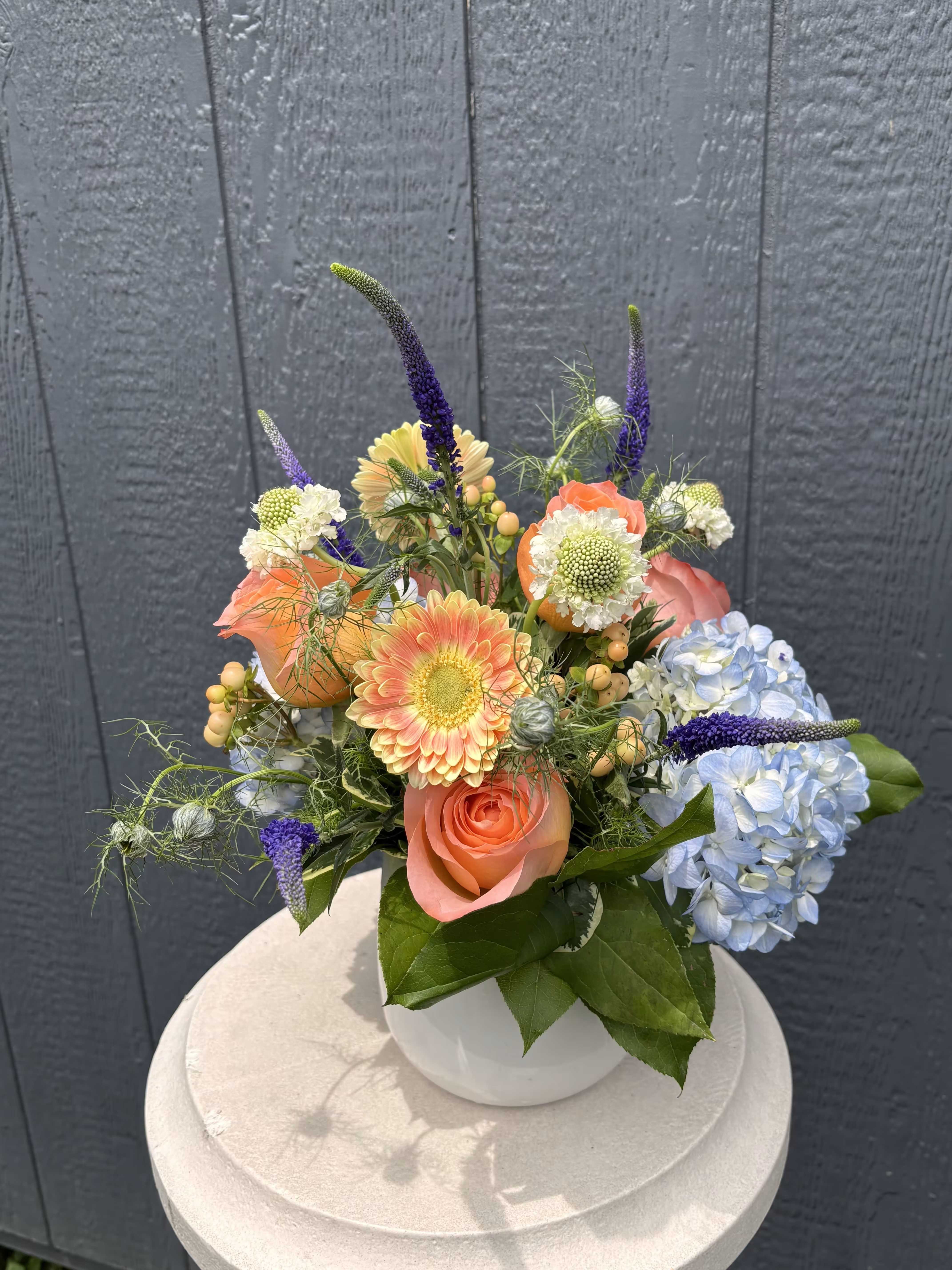 Blue Skies &amp; Blossoms - Like a perfect summer day, Blue Skies &amp; Blossoms brings together soft blue hydrangea, sunny peach gerbera daisies, coral roses, delicate scabiosa, and whimsical accents of veronica and seasonal textures. Designed in a classic white container, this cheerful arrangement feels fresh from the garden and full of summertime charm.  With its blend of soft pastels and vibrant blooms, Blue Skies &amp; Blossoms is perfect for birthdays, thank-yous, celebrations, or simply brightening someone's day. This garden-inspired design captures the beauty of long summer afternoons, blue skies, and flowers in full bloom.  Flowers and colors may vary slightly based on seasonal availability while maintaining the overall style and feel of the arrangement.