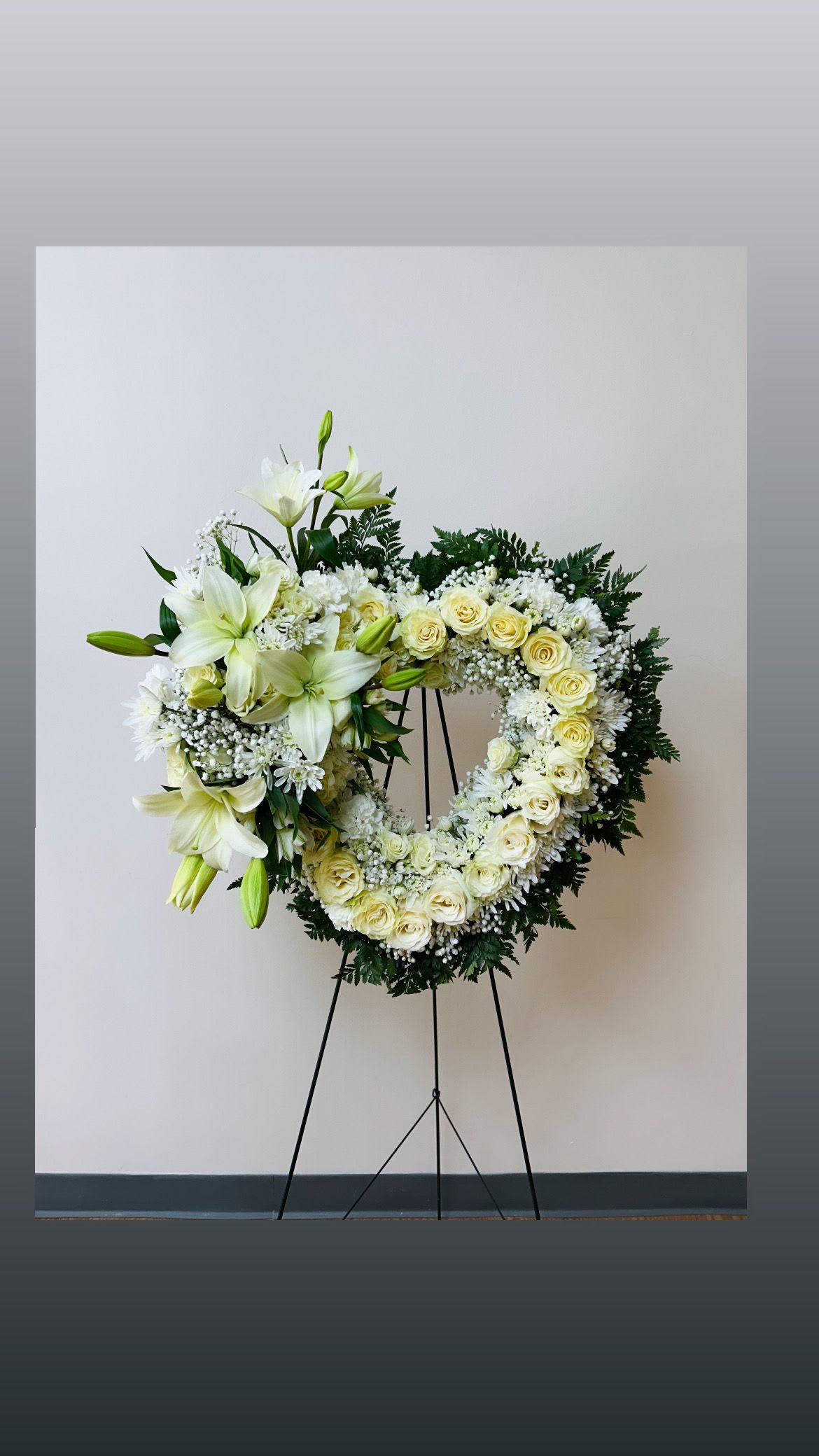 Heart-shaped white floral wreath on a stand with lilies and roses