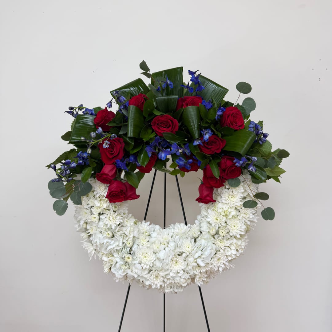 Red roses and blue flowers arranged in a white floral wreath on a stand